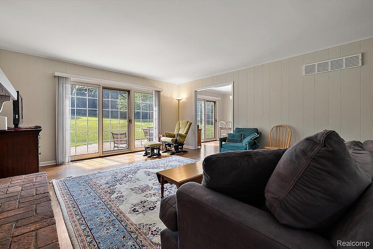 This is an interior shot of a living room featuring a large area rug, a fireplace, and sliding glass doors leading to the outside. The room is furnished with a sofa, a rocking chair, and a small table. The walls are paneled, and the overall style is traditional and comfortable.