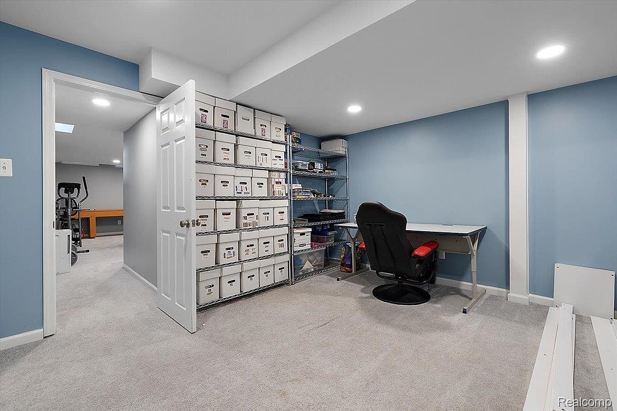 This is a well-organized basement space, featuring light blue walls and light gray carpet. The room includes a desk with a black office chair, metal shelving units filled with storage boxes, and an open doorway leading to another area with exercise equipment. The space appears clean and functional, suitable for an office or hobby area.