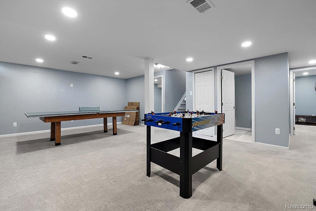 This is a spacious finished basement featuring neutral-toned carpet and light blue walls. The room includes a ping pong table, a foosball table, and multiple doorways, suggesting access to other areas. Recessed lighting provides ample illumination, creating a bright and inviting atmosphere.