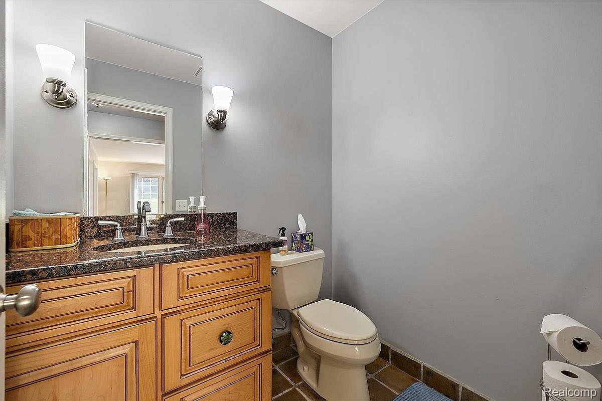 This is a bathroom featuring a vanity with a dark countertop and wooden cabinets. A mirror hangs above the sink, and there are sconce lights on either side. A toilet is positioned to the right, and the floor is tiled. The walls are painted a neutral gray color, creating a clean and functional space.