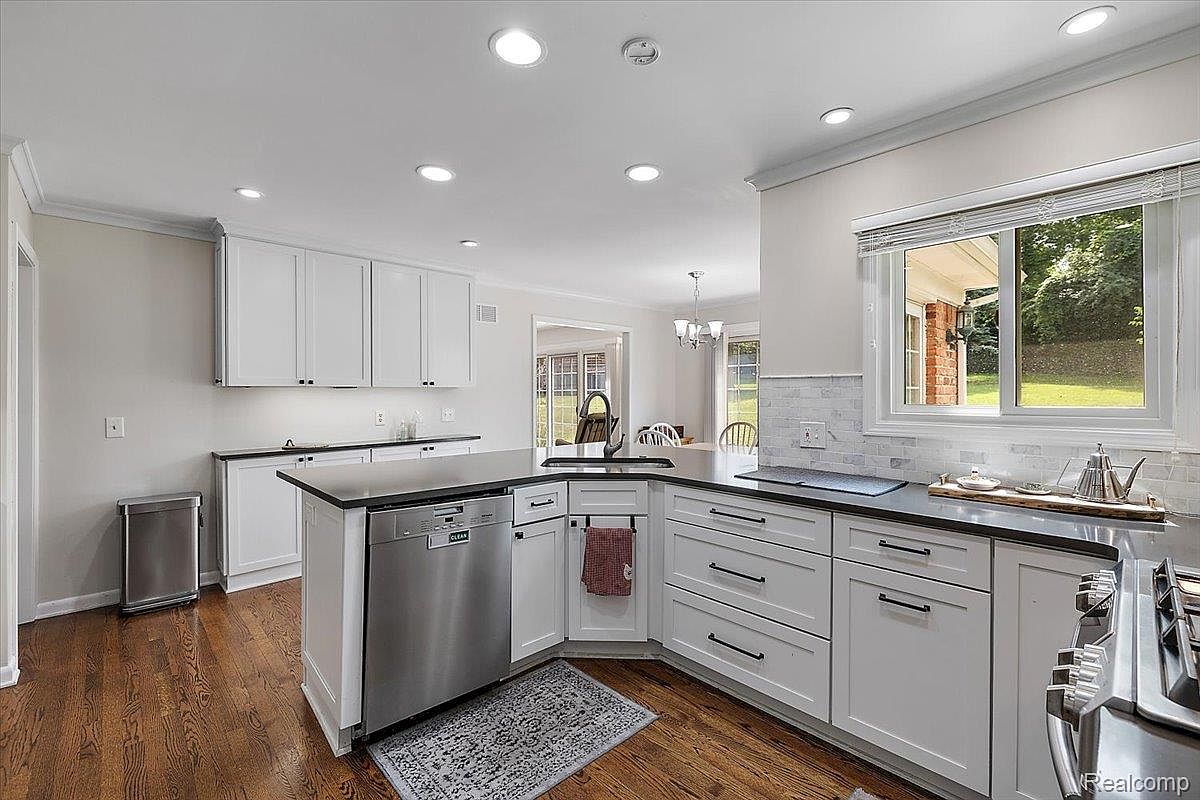 This is a well-lit kitchen featuring white cabinets, black countertops, and stainless steel appliances, including a dishwasher. The hardwood floors add warmth to the space, and a window provides natural light and a view of the outdoors. The kitchen has a clean and modern aesthetic.