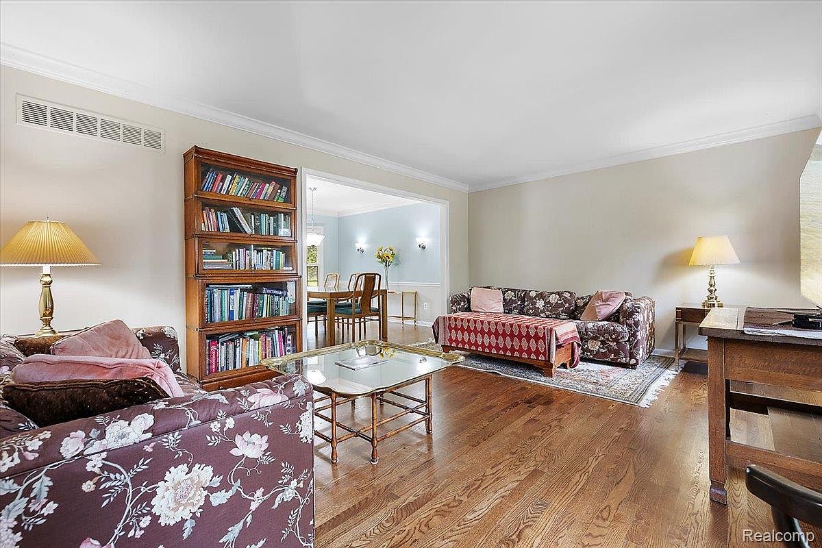 This living room features hardwood floors, floral patterned sofas, and a wooden bookshelf filled with books. A glass-topped coffee table sits in the center of the room, and a doorway leads to the dining area. The room has a cozy and inviting atmosphere.