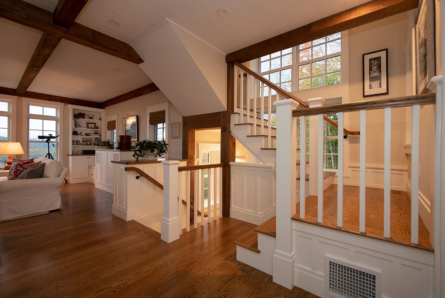 This interior view captures a sophisticated staircase area featuring warm wood flooring and exposed timber ceiling beams. The space is defined by white wainscoting, a classic white balustrade, and a landing that leads to a bright, window-filled stairwell. To the left, a glimpse of a cozy living area with built-in shelving and a telescope creates an inviting, open-concept atmosphere.