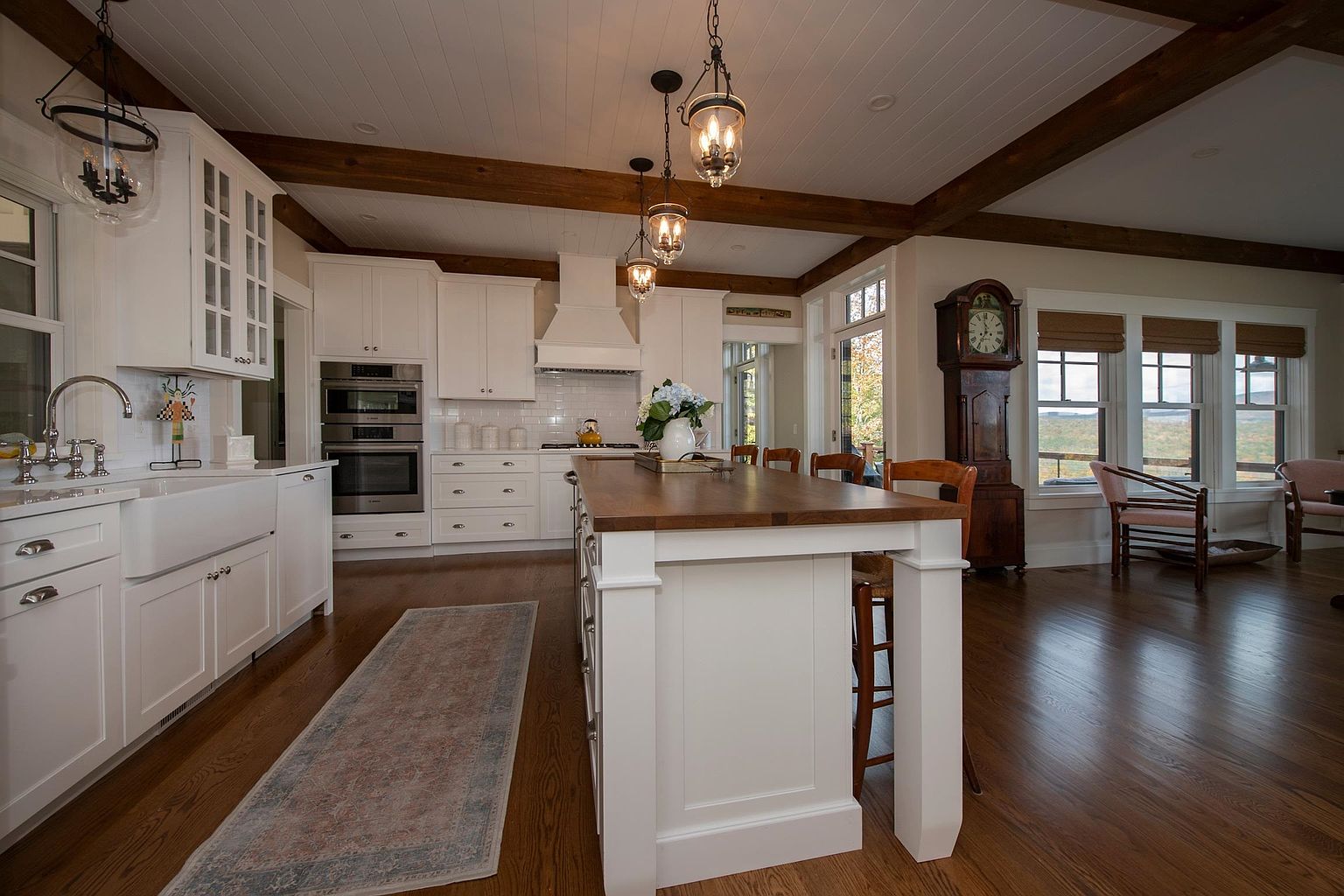 This bright and spacious kitchen features white cabinetry, a large central island with a dark wood countertop, and warm-toned hardwood flooring. The space is accented by exposed wooden ceiling beams and elegant pendant lighting, while a tall grandfather clock adds a touch of traditional charm. Large windows provide natural light and a view of the outdoors, creating an inviting and well-appointed atmosphere.