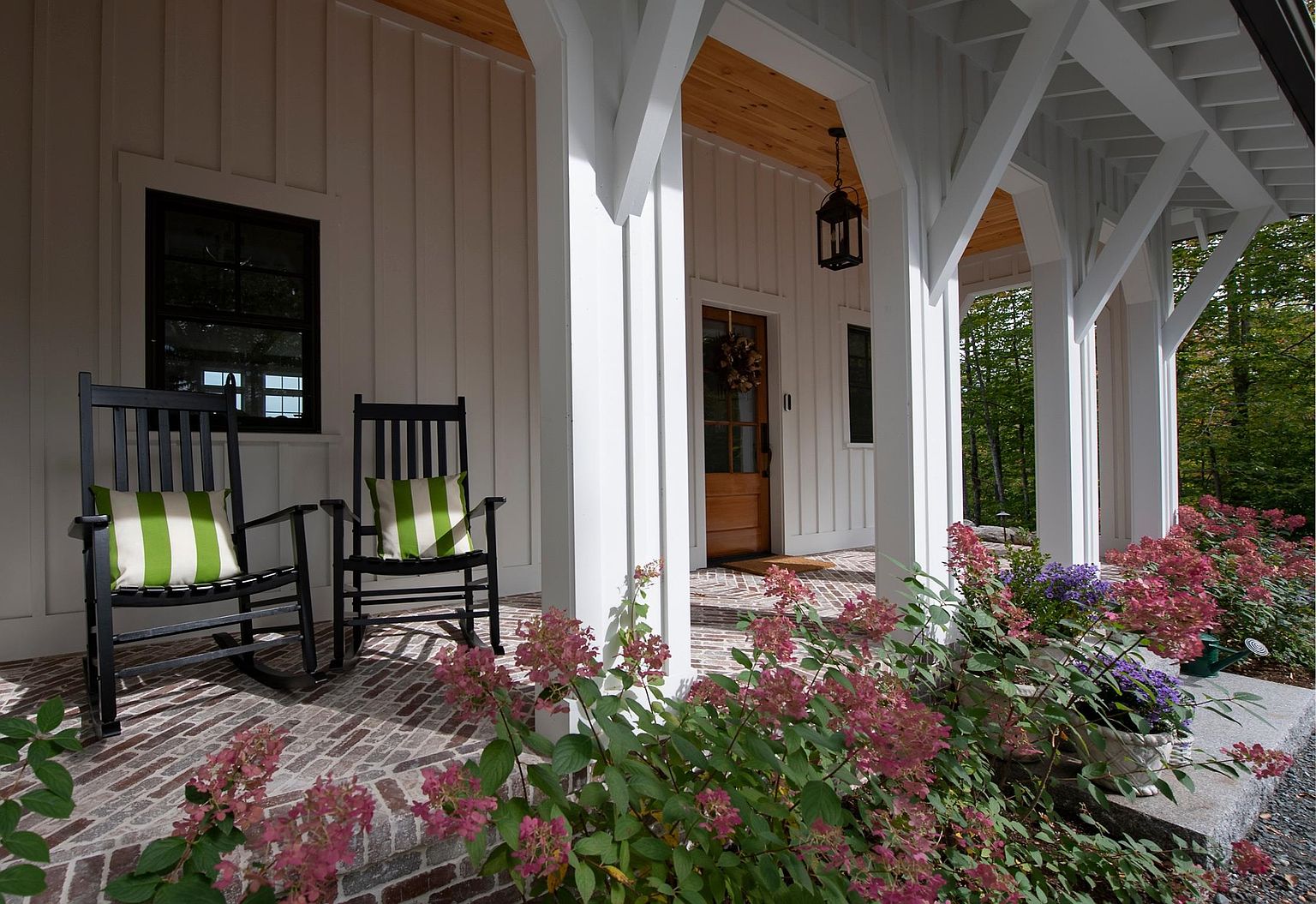 This inviting front porch features a classic farmhouse aesthetic with white board-and-batten siding and elegant arched architectural supports. Two black rocking chairs with green-and-white striped cushions sit on a herringbone-patterned brick floor, while lush pink hydrangeas frame the foreground. The warm wood front door and natural wood ceiling accents create a welcoming, high-end curb appeal.