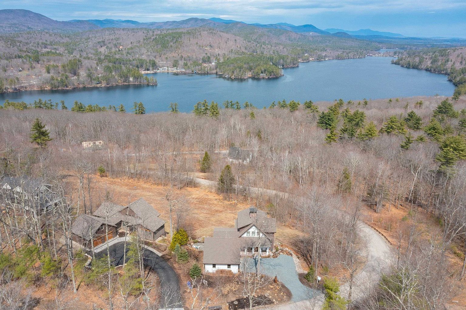 This high-angle aerial shot captures a secluded residential property nestled within a dense, wooded landscape overlooking a large, serene lake. The scene features two distinct multi-story homes with dark-toned roofs, each with its own driveway, surrounded by a mix of evergreen and deciduous trees. The expansive view highlights the property's tranquil setting, emphasizing its connection to nature and the surrounding mountainous terrain.