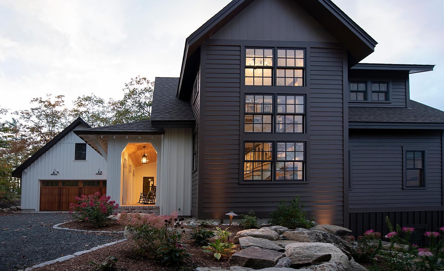 This striking modern farmhouse features a high-contrast exterior with dark horizontal siding paired with white vertical board-and-batten accents. A prominent, multi-pane window stack draws the eye, while a welcoming covered entryway with warm wood-toned ceiling details creates an inviting focal point. The property is set against a natural landscape with a gravel driveway, stone landscaping, and mature trees, offering a sophisticated and cozy curb appeal.