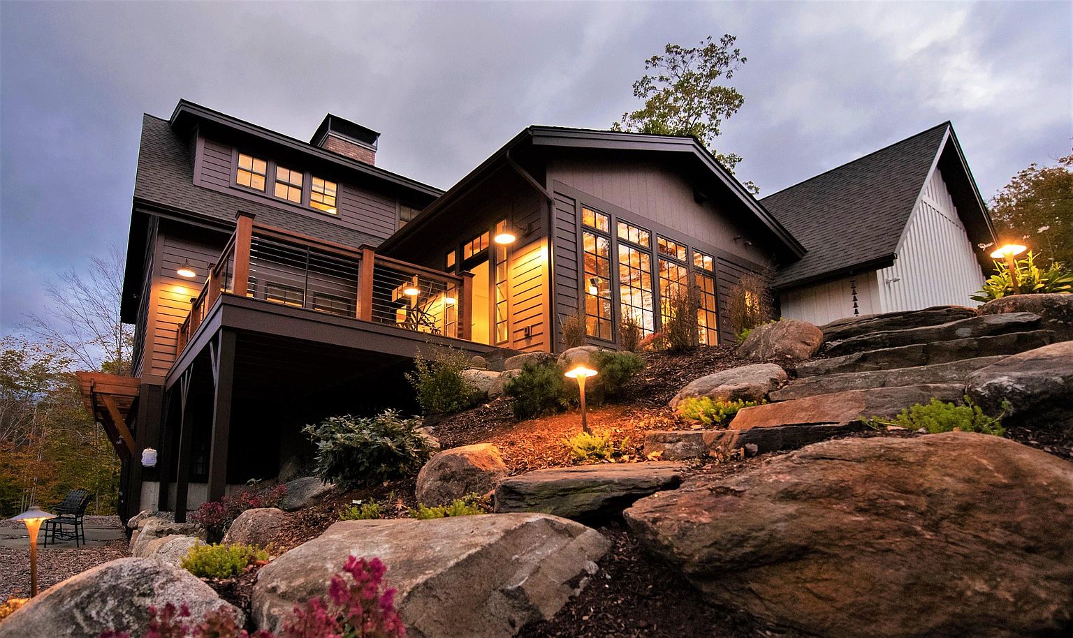 This striking rear exterior view captures a modern home at dusk, featuring dark-toned horizontal siding contrasted by a bright white vertical-board section. A spacious elevated deck with a metal railing extends from the main structure, while a naturalistic stone staircase winds through a landscaped rock garden in the foreground. Warm exterior lighting illuminates the home's windows and the path, creating an inviting and sophisticated atmosphere.