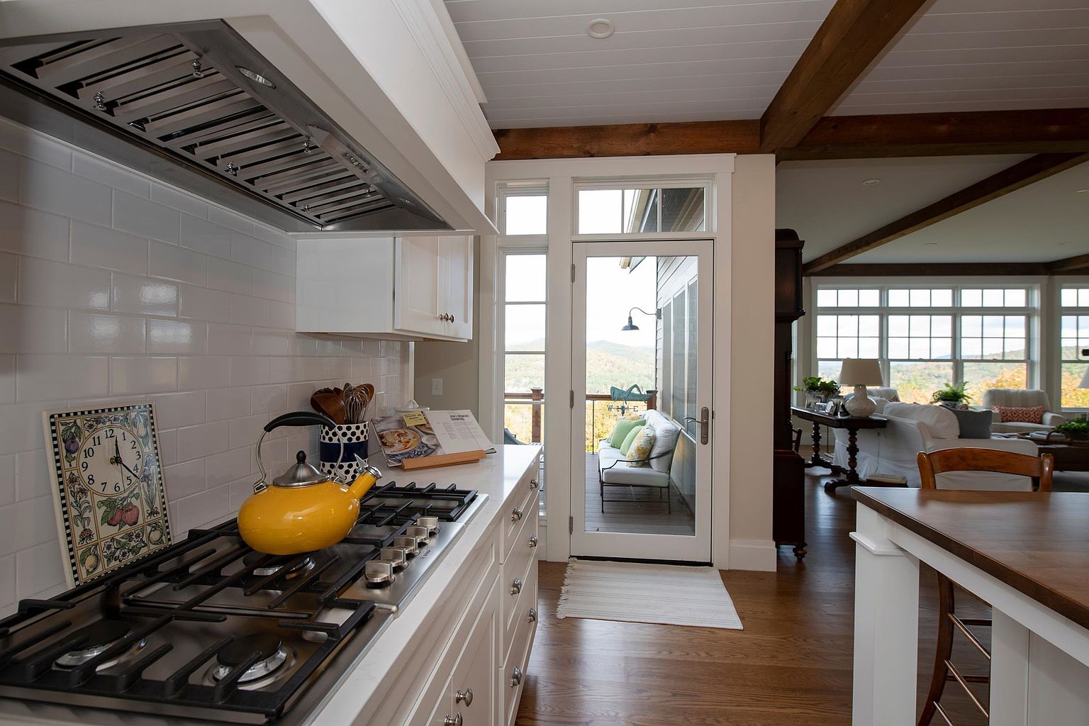 This bright kitchen features white cabinetry, a subway tile backsplash, and a stainless steel gas range topped with a vibrant yellow kettle. A glass door provides a seamless transition to an outdoor deck, while the open floor plan reveals a glimpse of a cozy living area with wood-beamed ceilings. The perspective captures the functional workspace of the kitchen while highlighting the home's connection to the scenic mountain views outside.