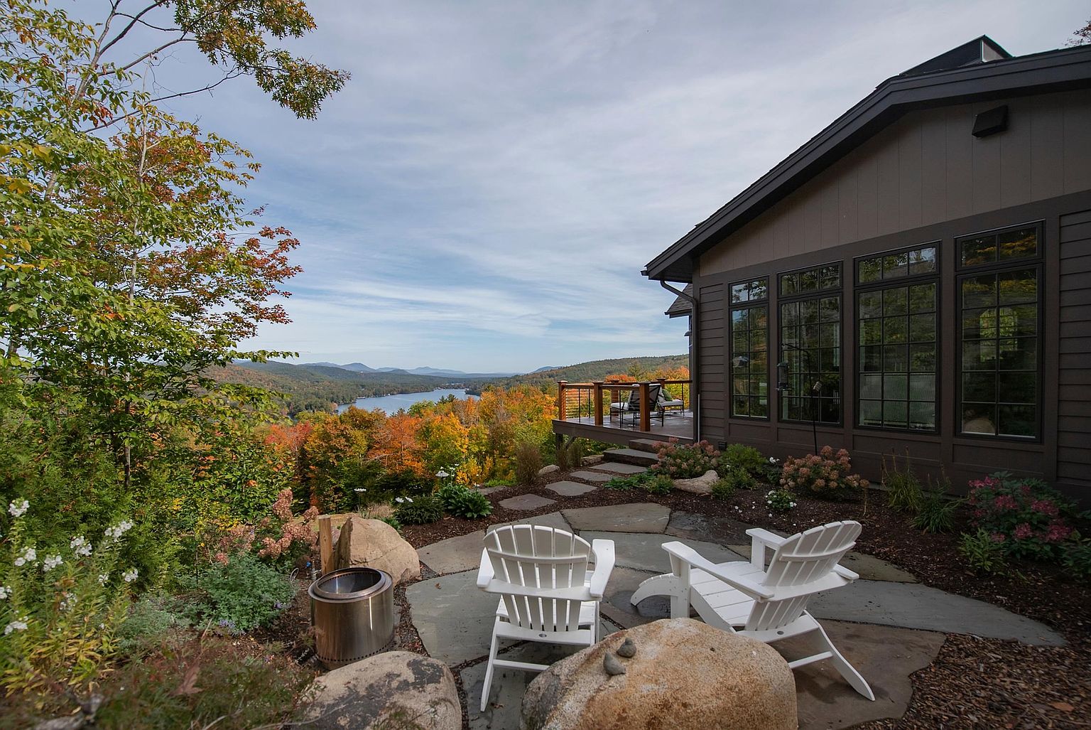 This scenic outdoor living space features a stone patio furnished with two white Adirondack chairs and a stainless steel fire pit, set against a backdrop of lush autumn foliage and a distant lake. A raised wooden deck extends from the side of the dark-toned home, which is accented by large windows that reflect the surrounding landscape. The perspective captures a tranquil, elevated vantage point that emphasizes the property's integration with its natural, mountainous environment.