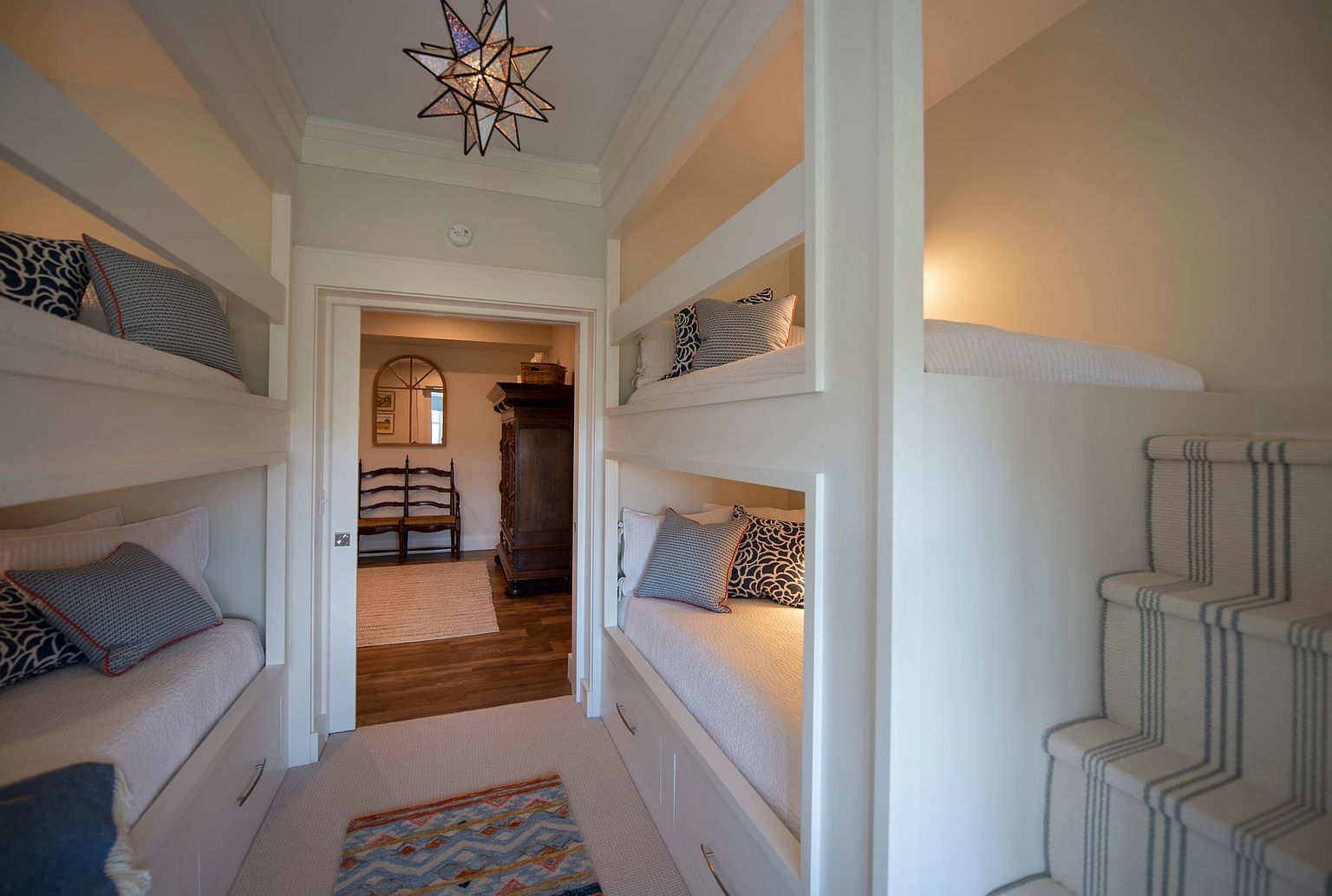This cozy bunk room features built-in sleeping nooks with integrated storage drawers and patterned bedding, maximizing space efficiency. A central hallway leads through an open doorway into an adjacent room, while a carpeted staircase with striped runners provides access to an upper bunk. The space is illuminated by a distinctive star-shaped pendant light, creating a charming and functional retreat.