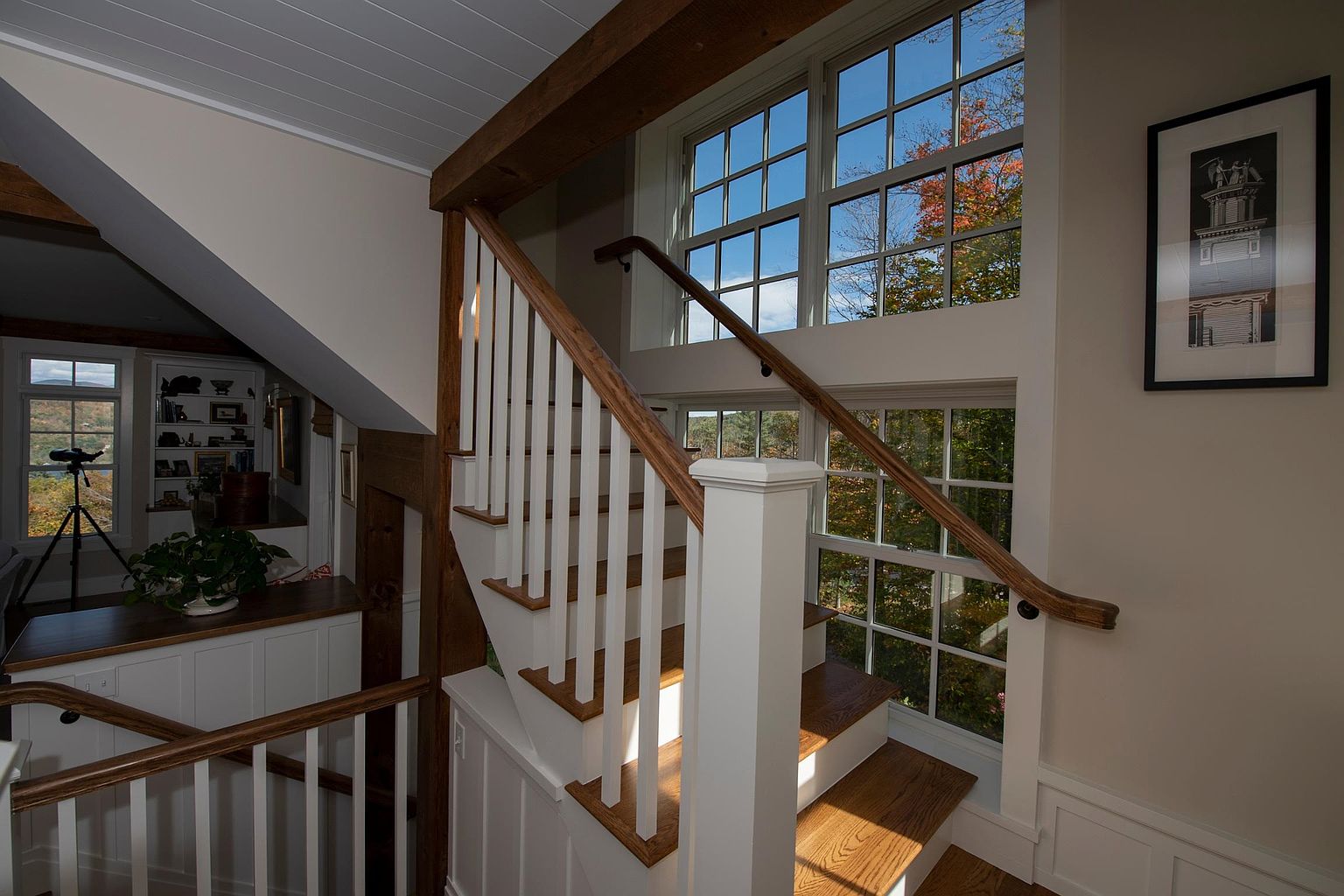 This interior view captures a staircase with white balusters and a warm-toned wood handrail, leading up toward a landing bathed in natural light. Large, multi-pane windows provide an expansive view of the surrounding autumn foliage, while a framed architectural print hangs on the adjacent wall. The space features exposed wood beams and a clean, traditional aesthetic that emphasizes the home's connection to the outdoors.