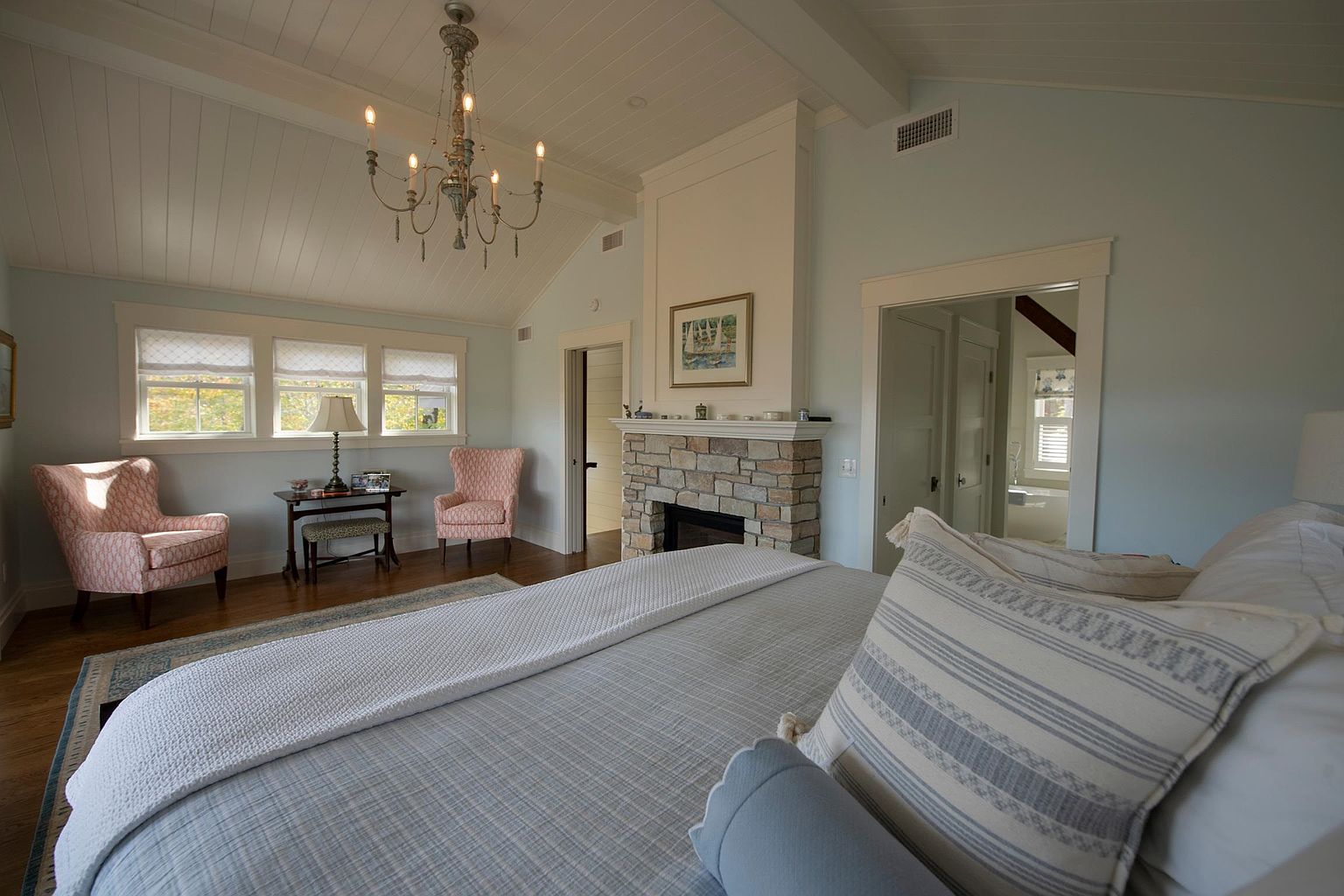 This spacious primary bedroom features a vaulted ceiling with white paneling and a central chandelier, creating an airy and elegant atmosphere. A stone-clad chimney breast serves as a focal point between two doorways, while a cozy seating area with two patterned armchairs sits near the windows. The room is anchored by a large bed with layered bedding, offering a comfortable and inviting retreat.