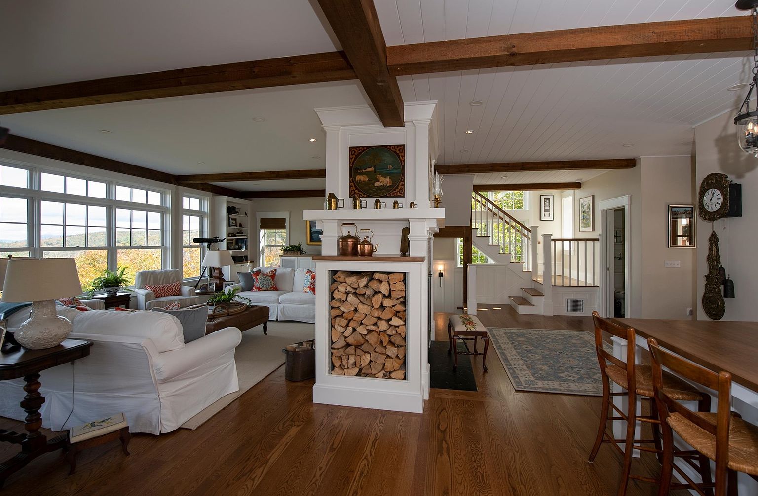 This open-concept living space features warm wood flooring, exposed ceiling beams, and a central fireplace structure with a built-in wood storage compartment. The room is bathed in natural light from large windows that offer scenic views, while a staircase leads to an upper level in the background. The decor is comfortable and inviting, blending traditional elements like a wall-mounted clock with a cozy, airy atmosphere.