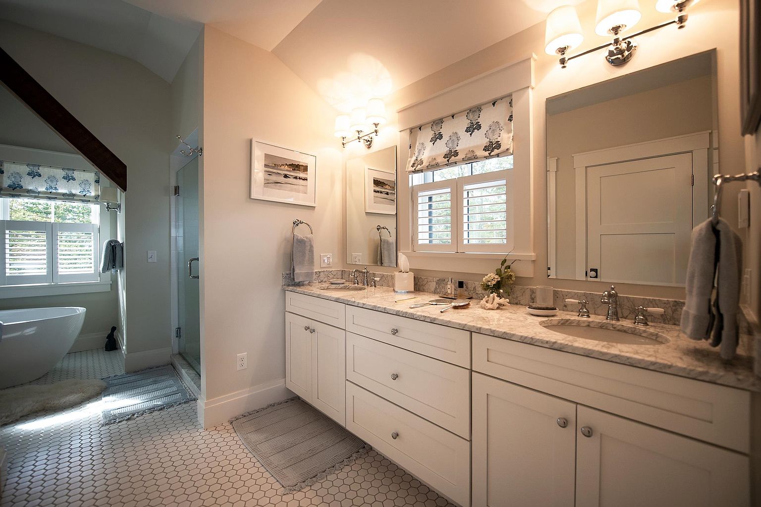 This bright and airy primary bathroom features a double vanity with a light-toned stone countertop and white cabinetry. To the left, a freestanding soaking tub sits near a window with plantation shutters, while a glass-enclosed shower is positioned between the tub and the vanity. The space is finished with small hexagonal floor tiles and classic wall sconces, creating a clean, sophisticated, and tranquil atmosphere.