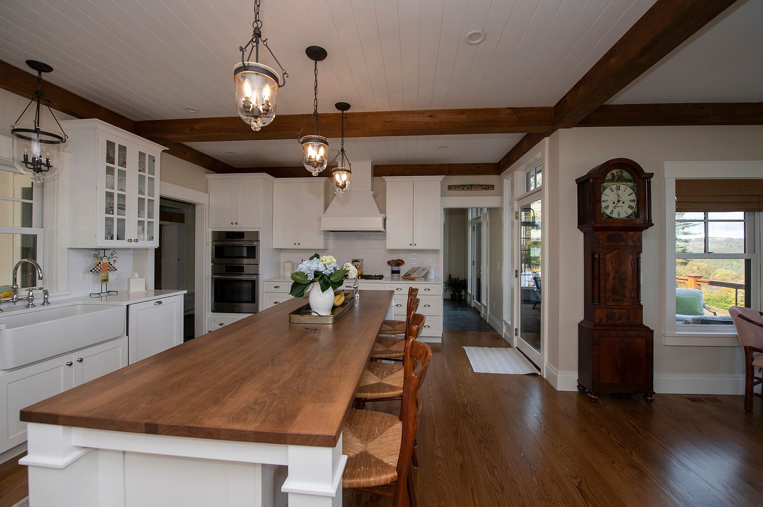 This bright and airy kitchen features a long central island with a wood-toned countertop and woven-seat bar stools, serving as the focal point of the room. White cabinetry, a farmhouse-style sink, and exposed ceiling beams create a classic, farmhouse-inspired aesthetic. A tall grandfather clock stands near a window that provides a view of the outdoors, while pendant lights hang above the island to complete the warm, inviting atmosphere.