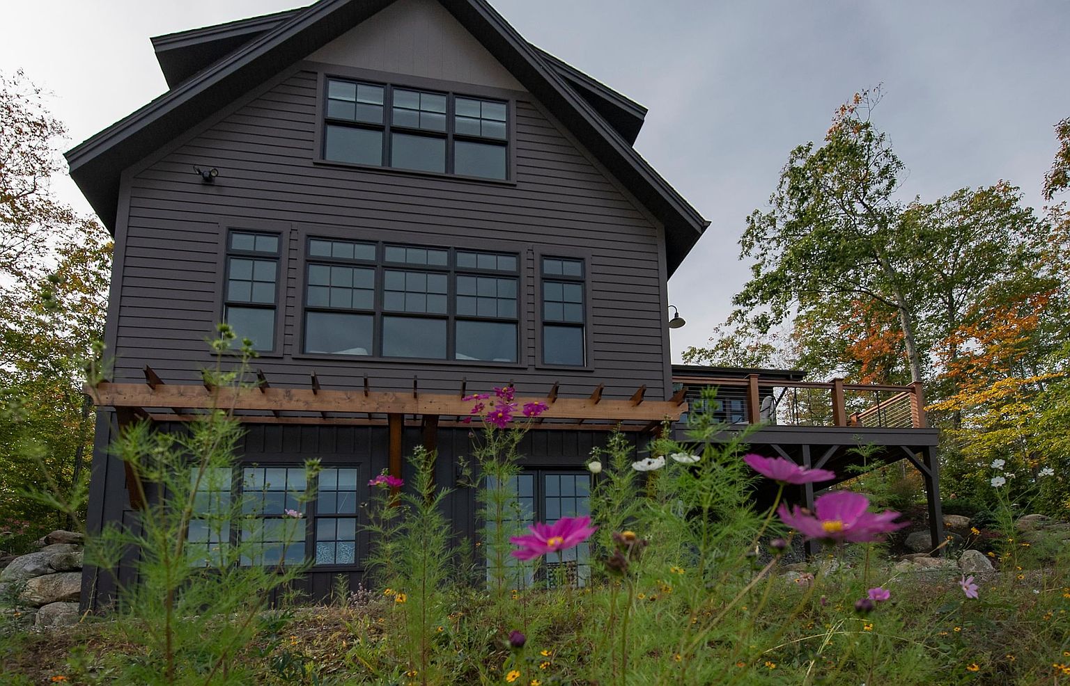 This rear view of a modern, dark-sided home showcases a multi-level design with a wooden pergola extending over the lower level and a raised deck on the right. Large, multi-pane windows dominate the facade, allowing for abundant natural light, while a foreground of vibrant wildflowers and greenery adds a soft, natural touch to the scene. The composition is captured from a low angle, emphasizing the home's architectural scale against a cloudy sky and autumn-toned trees.