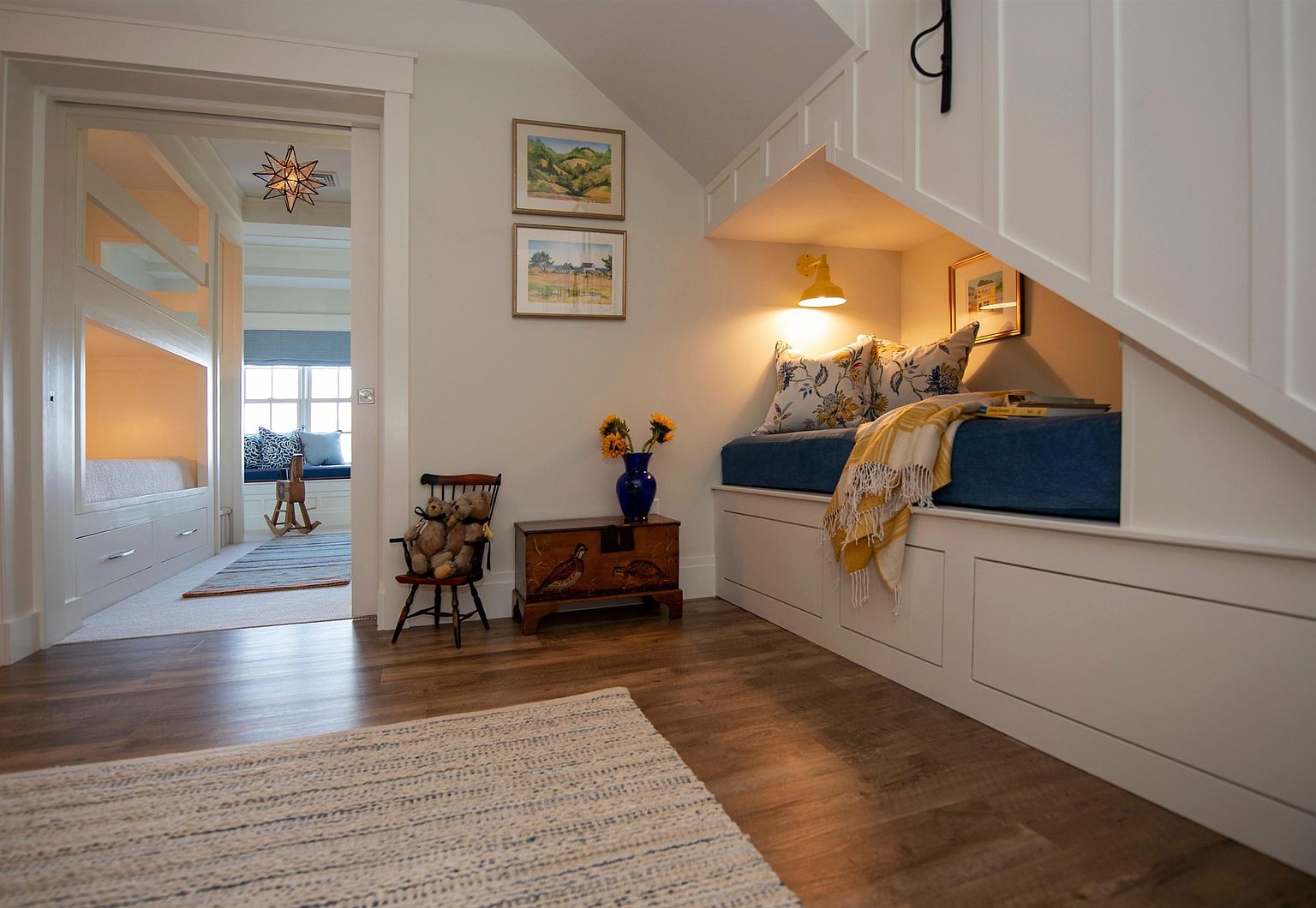 This cozy, well-lit hallway features a built-in reading nook tucked under a staircase, complete with a blue cushioned bench, patterned throw pillows, and a warm wall-mounted lamp. To the left, a doorway leads into a bright bedroom area with bunk beds, while a small wooden chest and a child-sized chair with teddy bears add a charming, lived-in touch. The space is finished with light-toned wood flooring and neutral walls, creating a welcoming and functional transition area.