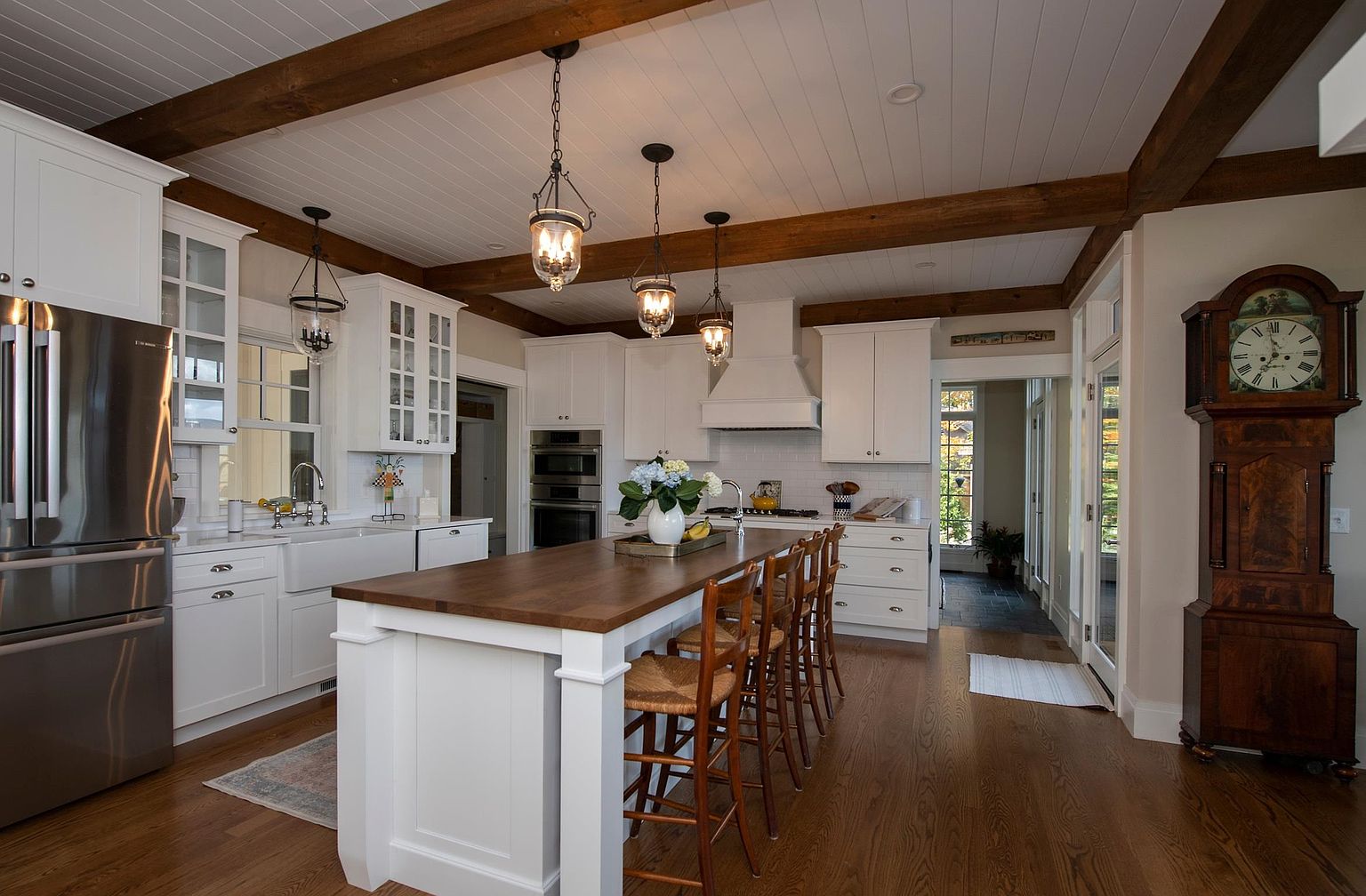 This bright and inviting kitchen features a large central island with a dark wood countertop and seating for four, serving as the focal point of the room. The space is defined by white cabinetry, a white subway tile backsplash, and exposed wooden ceiling beams that add a rustic touch to the clean, classic aesthetic. A tall, antique-style grandfather clock stands to the right, while a doorway leads to a sunlit hallway, creating a warm and well-appointed atmosphere.