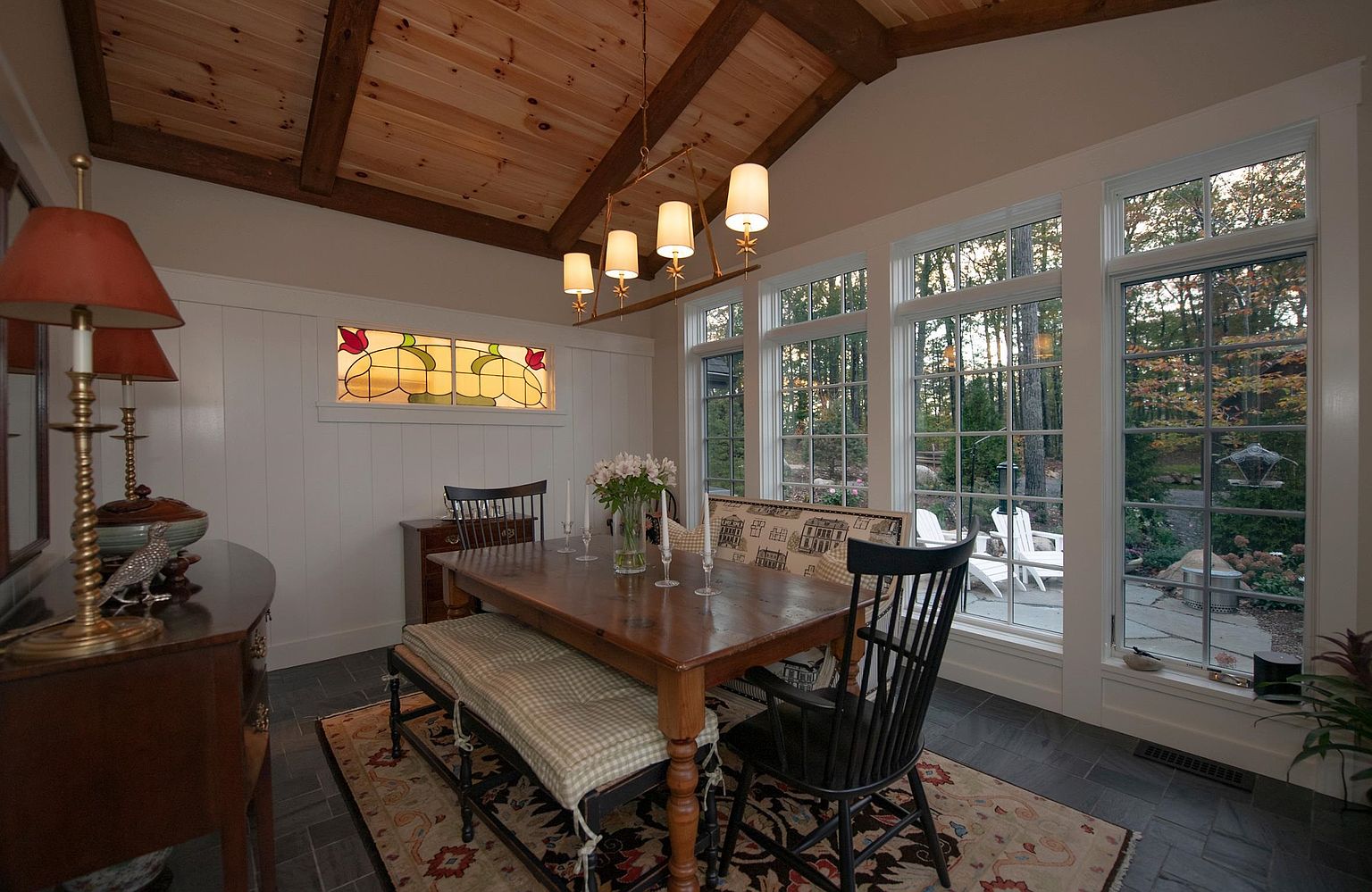 This cozy dining room features a rustic aesthetic with a wood-paneled vaulted ceiling, exposed beams, and dark stone flooring. A wooden dining table is centered on a patterned area rug, flanked by a bench with a gingham cushion and black spindle-back chairs. Large windows provide ample natural light and a view of the wooded backyard, while a stained-glass accent window and a multi-light chandelier add character to the space.