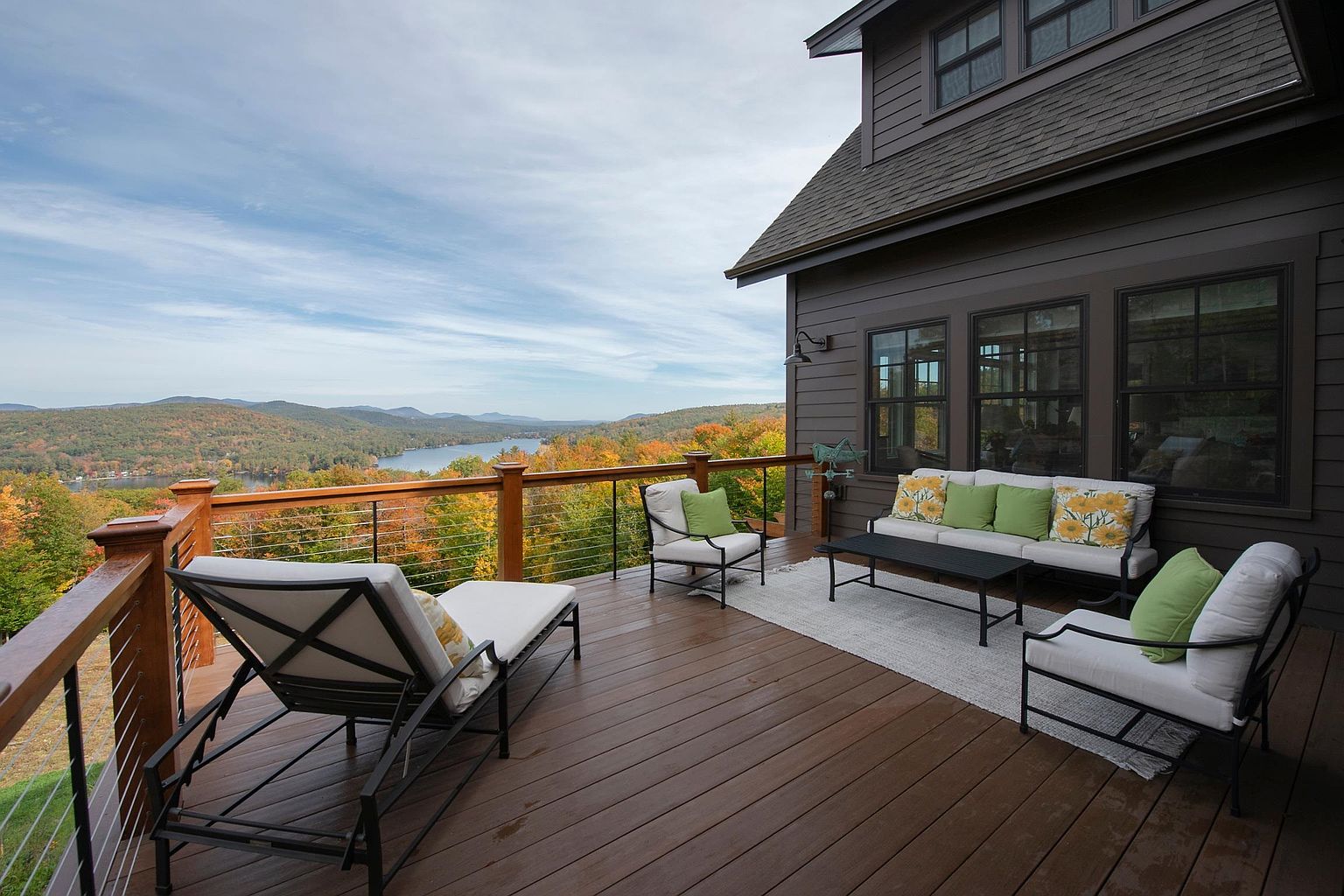 This expansive elevated deck offers a stunning panoramic view of a lake surrounded by autumn-colored forests and distant mountains. The space is furnished with a comfortable outdoor sofa, two armchairs, and a lounge chair, all arranged on a neutral area rug atop the dark wood-toned decking. A sleek cable railing system ensures unobstructed views, while the dark exterior siding of the home provides a sophisticated contrast to the vibrant natural landscape.