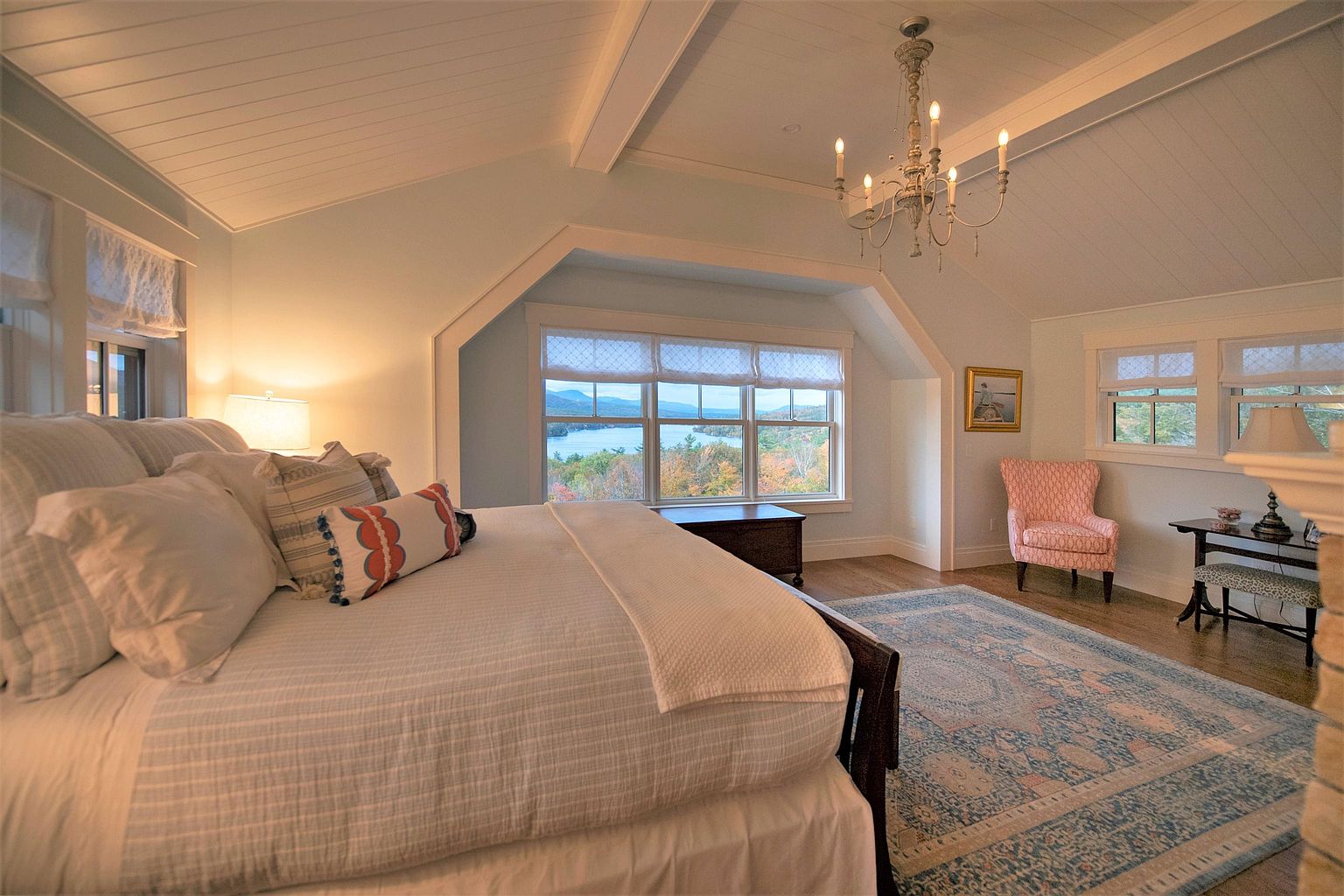 This spacious primary bedroom features a vaulted ceiling with white paneling and a classic chandelier, creating an airy and elegant atmosphere. A large bed with layered linens sits prominently in the room, positioned to take advantage of the expansive windows that offer a scenic view of the lake and surrounding trees. The space is complemented by a patterned area rug, a soft pink armchair, and a small writing desk, all contributing to a serene and comfortable retreat.
