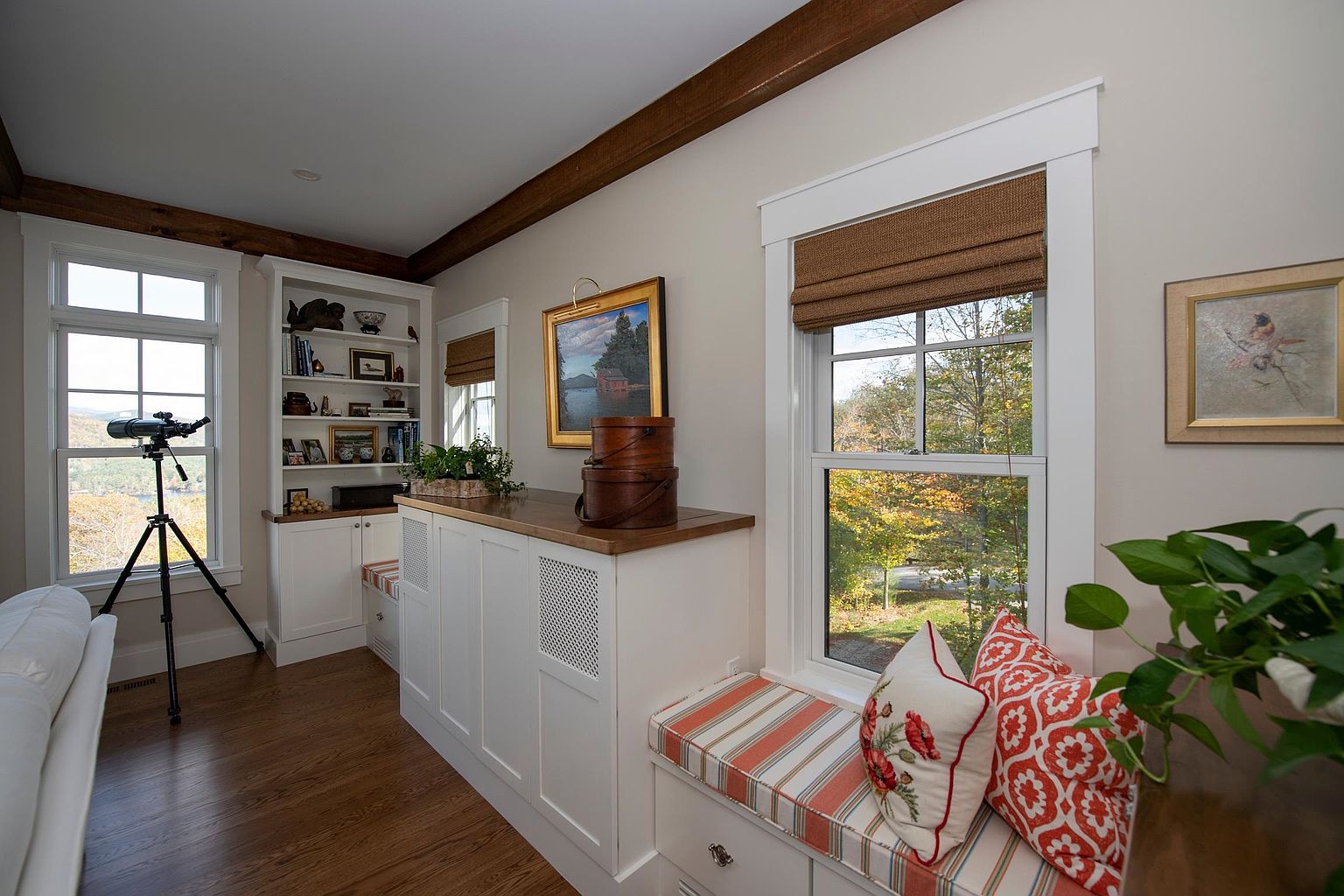 This cozy study features custom built-in cabinetry with open shelving and a window seat topped with patterned cushions. A telescope is positioned by the window, emphasizing the scenic outdoor view, while warm wood trim and beams add a rustic, inviting character to the space. The room is decorated with framed artwork and a wooden storage chest, creating a refined and tranquil atmosphere.