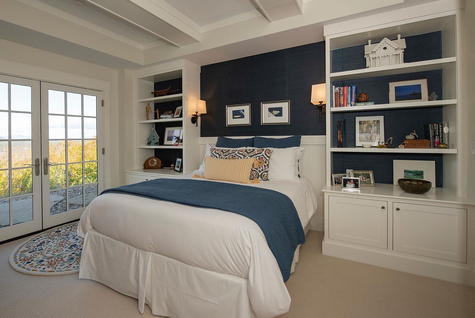 This cozy bedroom features a central bed dressed in white linens with a navy blue throw, flanked by symmetrical built-in shelving units against a dark blue accent wall. Large glass doors to the left provide natural light and a view of the outdoors, while the room is accented by a patterned area rug and wall-mounted sconces. The overall aesthetic is clean, organized, and inviting, with a balanced layout that maximizes storage and comfort.
