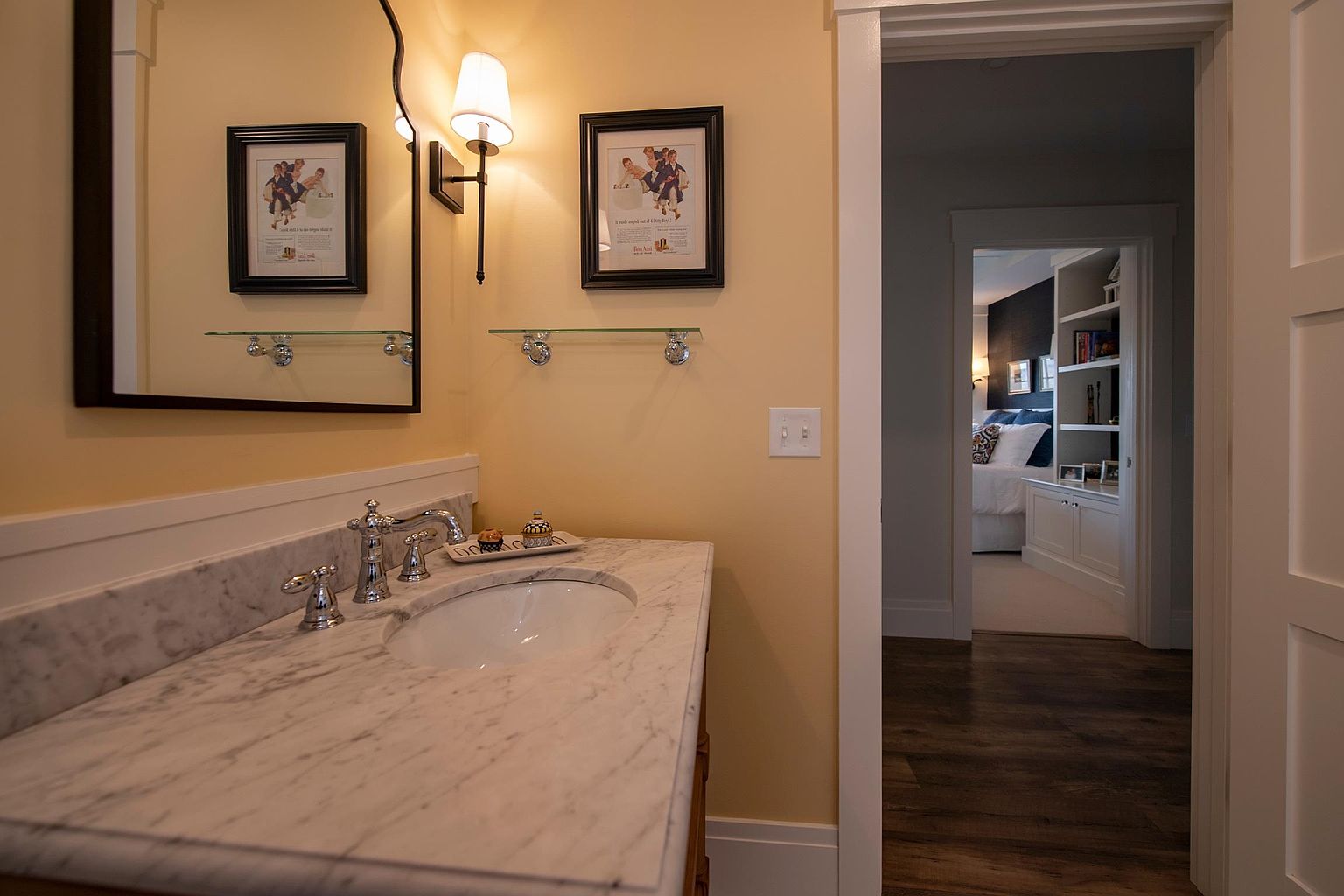 This bathroom features a vanity with a stone countertop and a classic faucet, set against warm-toned walls. A mirror hangs above the sink, flanked by framed artwork and a wall-mounted light fixture, while a glass shelf provides additional storage. The perspective looks through an open doorway into an adjacent bedroom, creating a sense of flow between the spaces.
