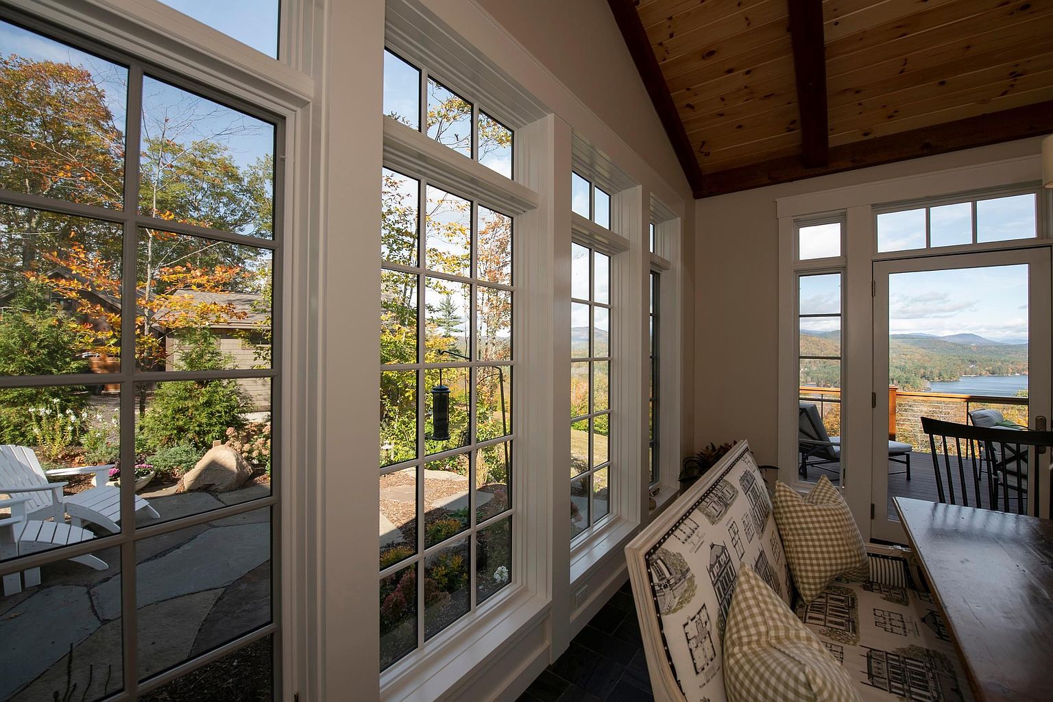 This bright sunroom features a series of tall, multi-pane windows that offer expansive views of the surrounding landscape and a distant lake. The space is furnished with a comfortable bench seat adorned with patterned cushions, set against a backdrop of a warm wood-paneled ceiling. The perspective captures the seamless transition between the cozy indoor seating area and the outdoor deck, emphasizing the home's connection to nature.
