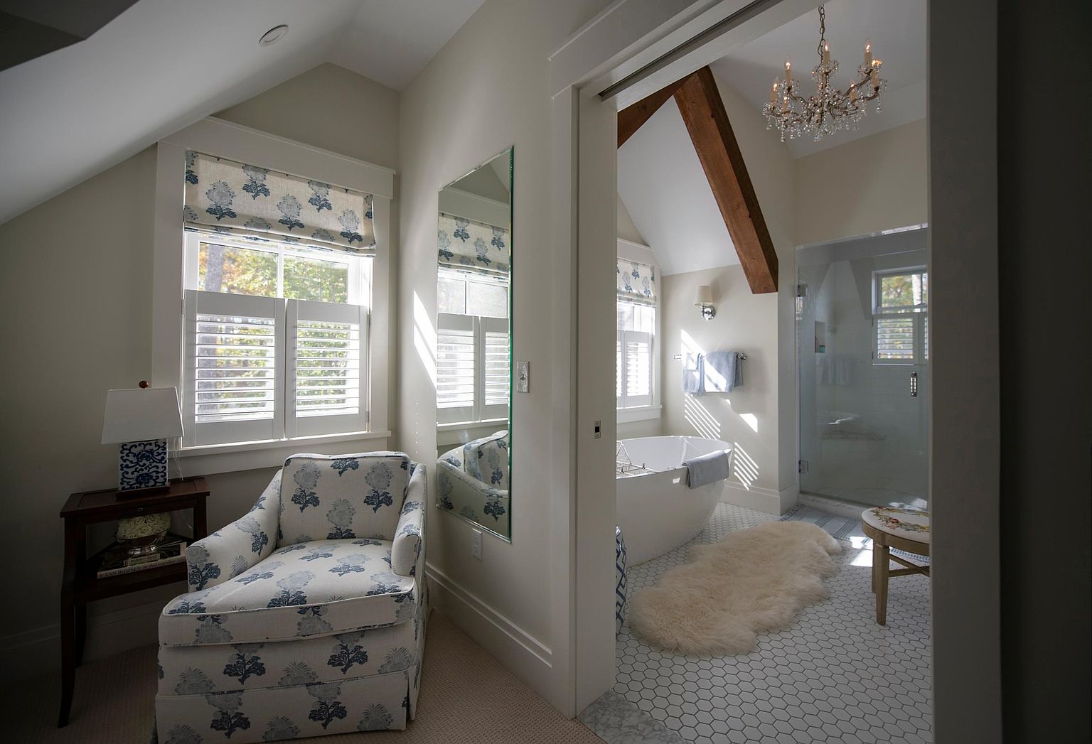 This elegant primary bathroom suite features a seamless transition from a cozy sitting area with a patterned armchair into a bright, spa-like bathing space. The bathroom is highlighted by a freestanding soaking tub, a glass-enclosed shower, and a striking exposed wooden beam overhead, all illuminated by a crystal chandelier. The space is finished with classic hexagonal floor tiles and soft, neutral wall tones, creating a serene and luxurious atmosphere.