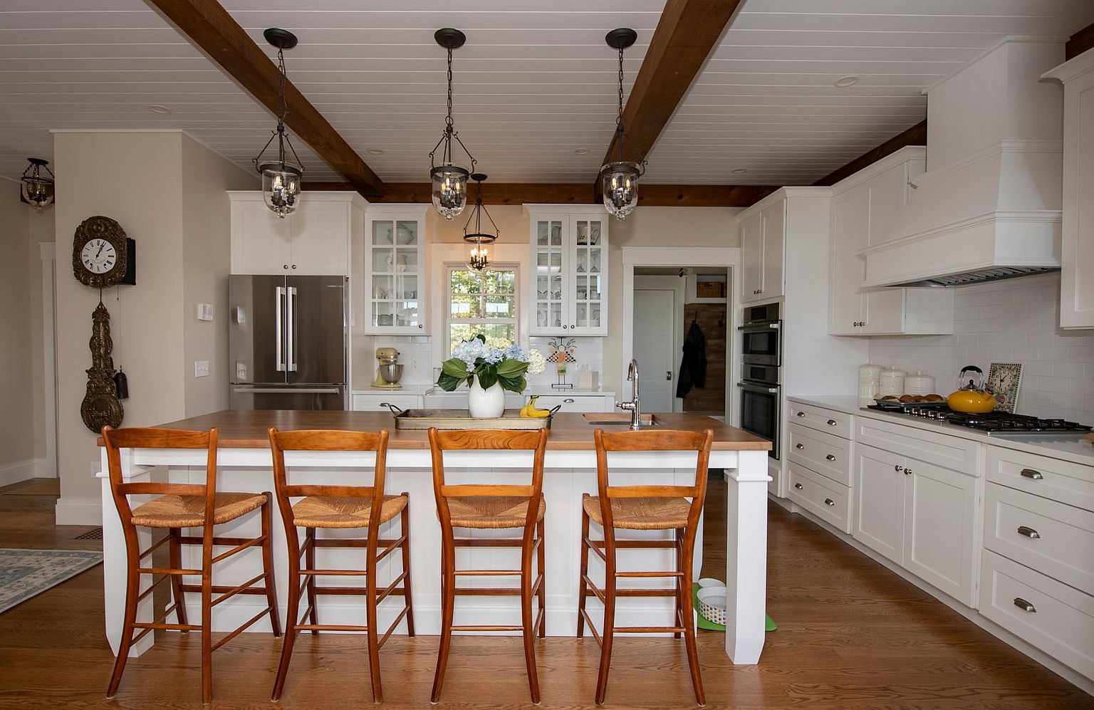 This bright and inviting kitchen features a large central island with a wood-toned countertop and four wooden bar stools, serving as the focal point of the room. The space is defined by white cabinetry, a subway tile backsplash, and exposed wooden ceiling beams that add a rustic touch to the clean, classic design. A window above the sink provides natural light, while three pendant lights hang above the island, creating a warm and functional atmosphere.