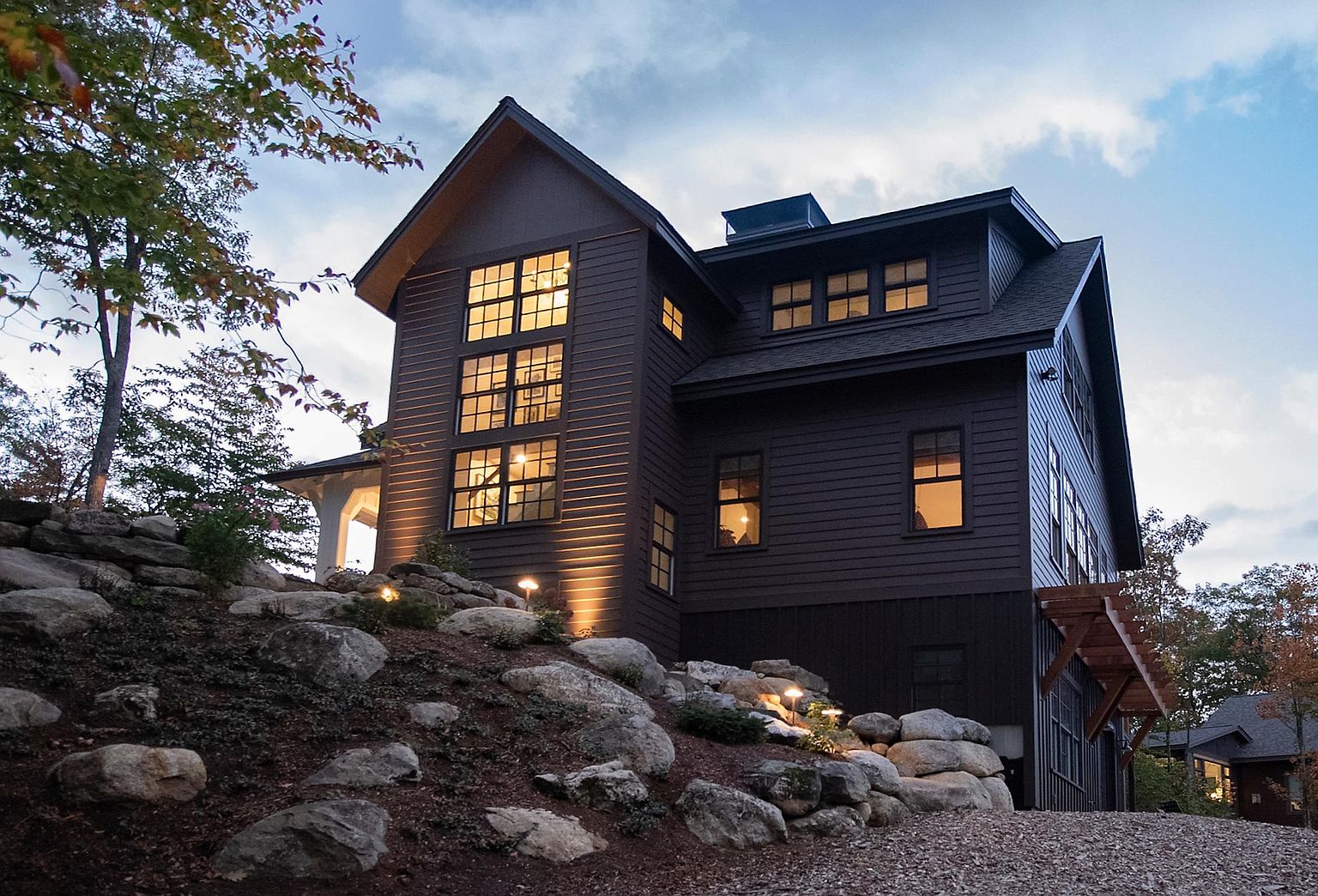 This striking multi-story home features dark horizontal siding and a prominent gable roofline, set against a rugged, landscaped hillside with large natural boulders. A dramatic vertical bank of windows illuminates the interior, casting a warm glow that contrasts with the twilight sky. The property is accented by subtle landscape lighting, highlighting the stone retaining walls and the textured exterior architecture.