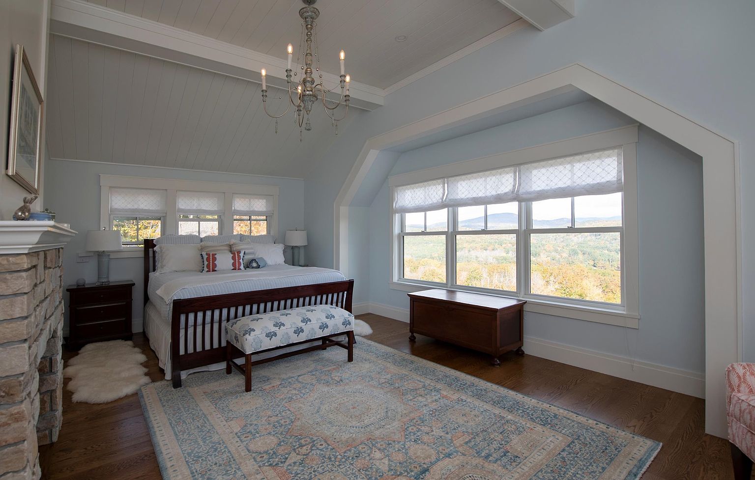 This spacious primary bedroom features a vaulted ceiling with exposed beams and a light blue color palette that creates a serene atmosphere. A dark wood bed frame is centered in the room, complemented by a patterned area rug and a stone-clad chimney breast rising above the mantel on the left. Large windows provide ample natural light and offer expansive views of the surrounding landscape, while a wooden chest sits at the foot of the window wall.