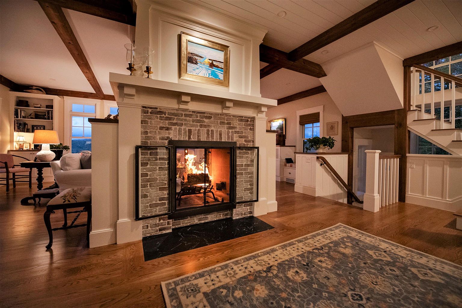 This inviting living room centers around a prominent brick fireplace with a dark stone hearth and a classic mantel topped with a framed painting. The space features warm wood flooring, exposed ceiling beams, and a neutral color palette that creates a cozy, rustic atmosphere. A staircase with white railings and wood accents leads to an upper level, while a patterned area rug anchors the seating area, providing a comfortable and well-appointed living space.
