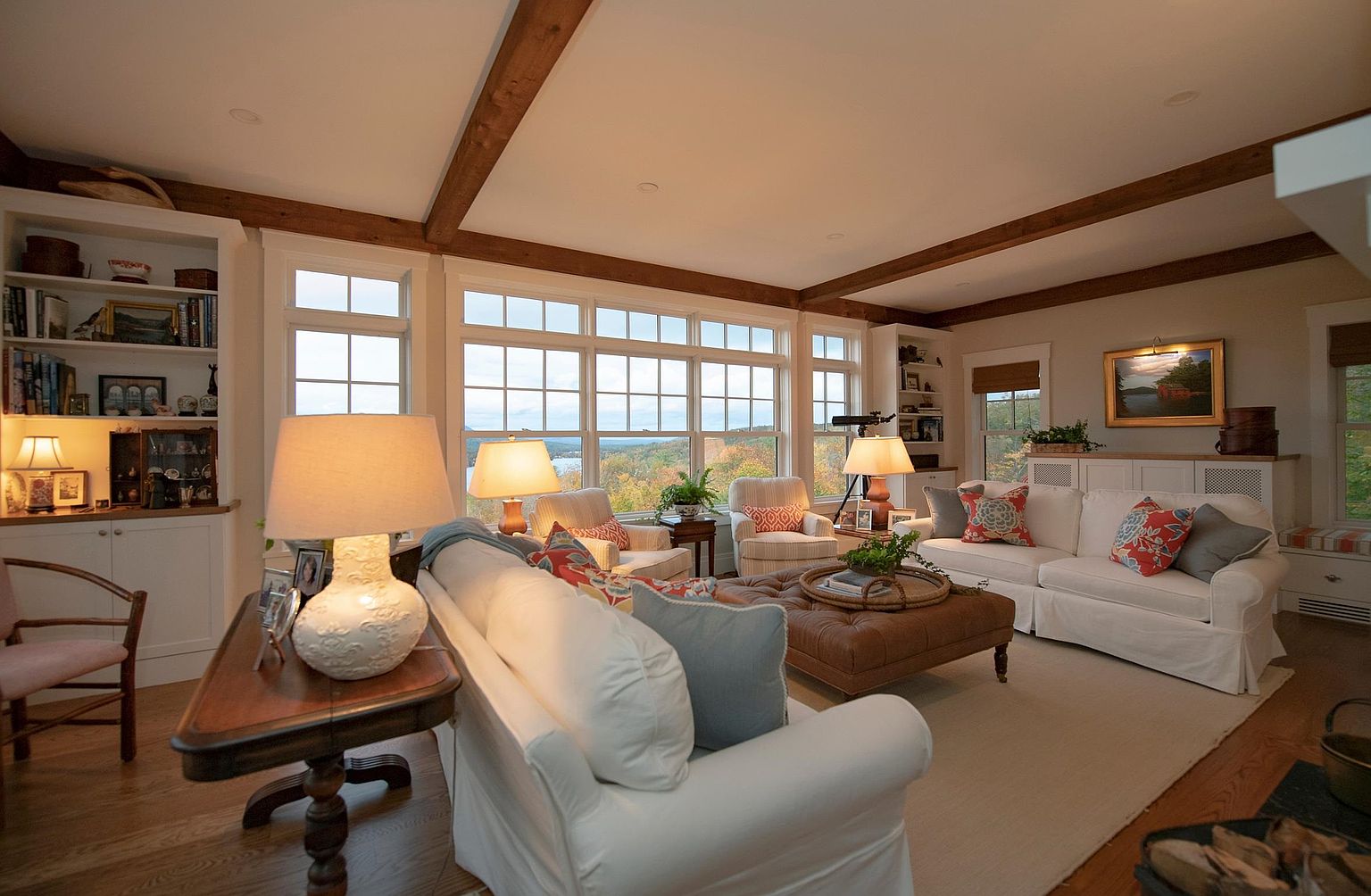 This inviting living room features a warm, traditional aesthetic with exposed wood ceiling beams and light-toned walls. The space is anchored by a large, plush white sofa and a leather ottoman, complemented by a pair of armchairs and a console table with a decorative lamp. Large windows provide an expansive view of the surrounding landscape, filling the room with natural light and creating a serene, comfortable atmosphere.