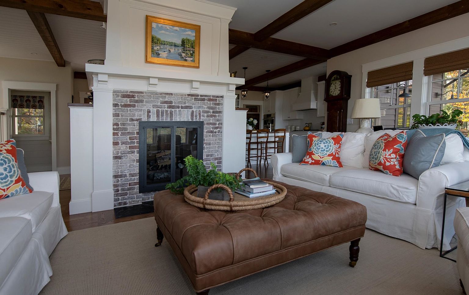 This inviting living room features a central brick fireplace with a white mantel and a framed painting above, serving as the focal point of the space. A large, tufted brown leather ottoman sits on a neutral area rug, surrounded by plush white sofas accented with colorful patterned pillows. The open-concept design allows for a clear view into the adjacent kitchen, while exposed dark wood ceiling beams add a touch of rustic character to the bright, airy room.
