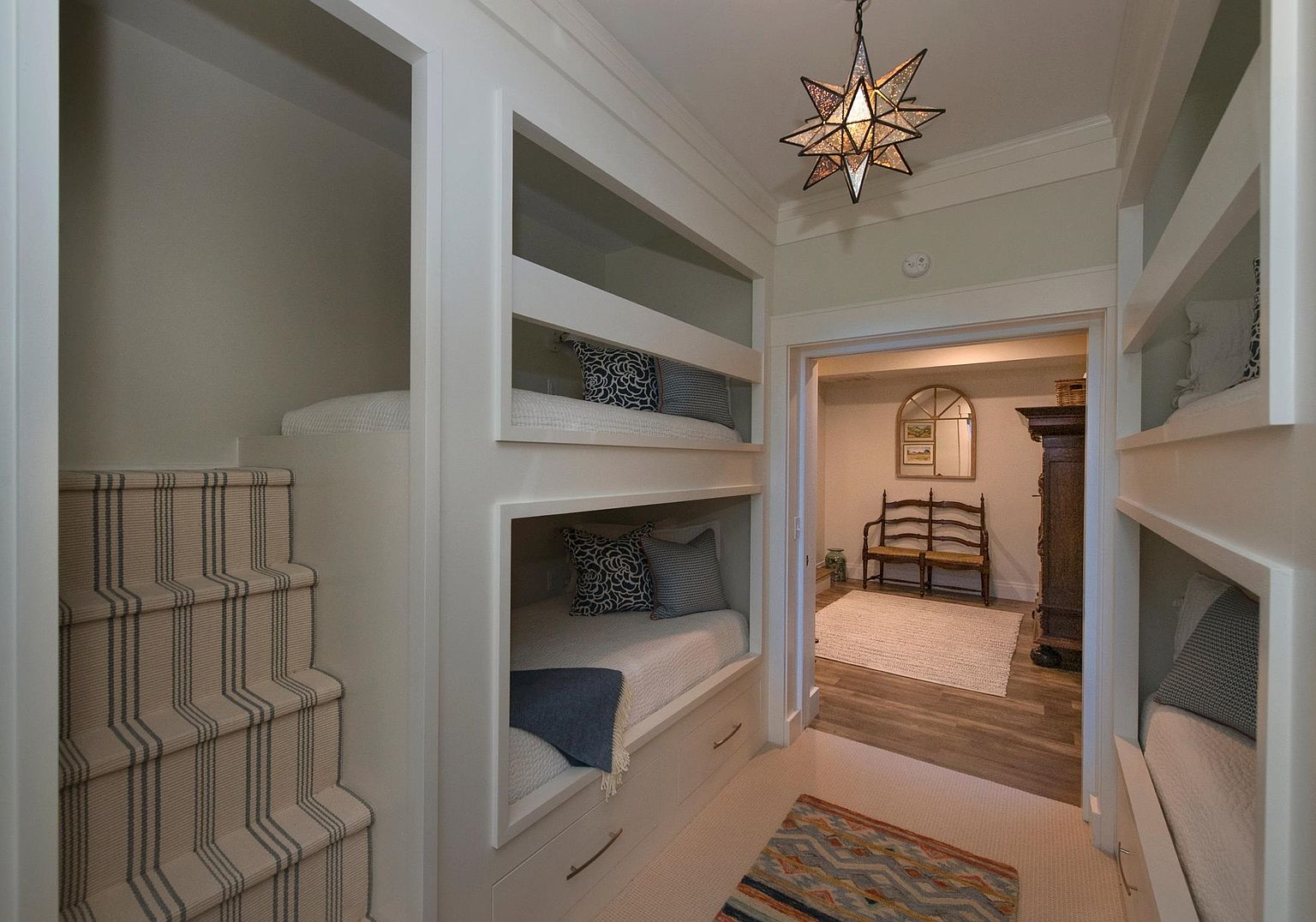 This unique bedroom space features built-in bunk beds on both sides of a central hallway, maximizing sleeping capacity with a custom, space-saving design. The room is accented by a distinctive star-shaped pendant light and a staircase with striped carpeting leading to an upper bunk. Through the doorway, a secondary room is visible, furnished with a wooden bench and a decorative mirror, creating a cohesive and inviting transition between spaces.