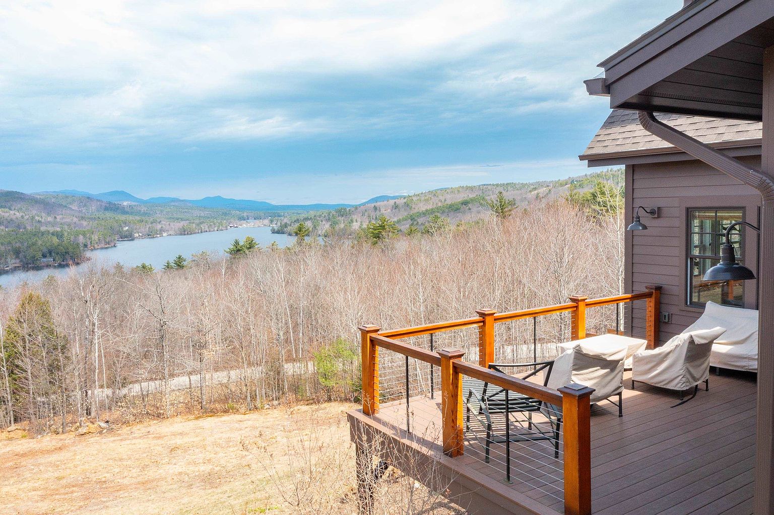 This elevated wooden deck offers a panoramic view of a serene lake surrounded by rolling, forested hills under a cloudy sky. The deck is furnished with comfortable outdoor seating covered in protective fabric, featuring a modern railing with thin metal cables. The perspective captures the seamless transition between the home's exterior architecture and the expansive natural landscape.