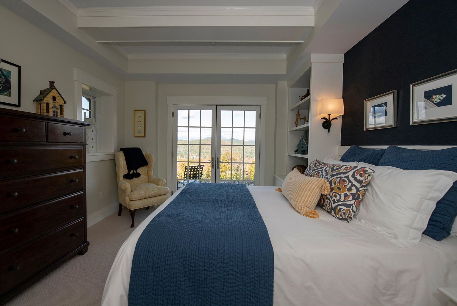 This inviting primary bedroom features a plush bed dressed in white linens with a deep blue quilted runner and patterned accent pillows. To the right, built-in shelving flanks a wall with a dark textured finish, while a cozy yellow armchair sits near a set of French doors that open to a scenic outdoor view. The room is accented by a dark wood dresser, framed artwork, and a warm wall-mounted lamp, creating a serene and well-appointed retreat.