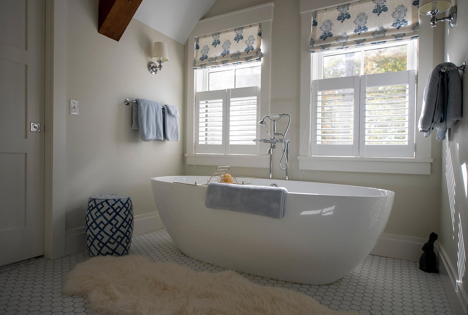 This serene primary bathroom features a freestanding soaking tub as the centerpiece, positioned beneath two windows adorned with white shutters and patterned valances. The space is finished with a white hexagonal tile floor, a plush faux-fur rug, and a decorative blue and white patterned stool. Chrome fixtures and soft blue towels add a refined, tranquil touch to this bright and airy retreat.