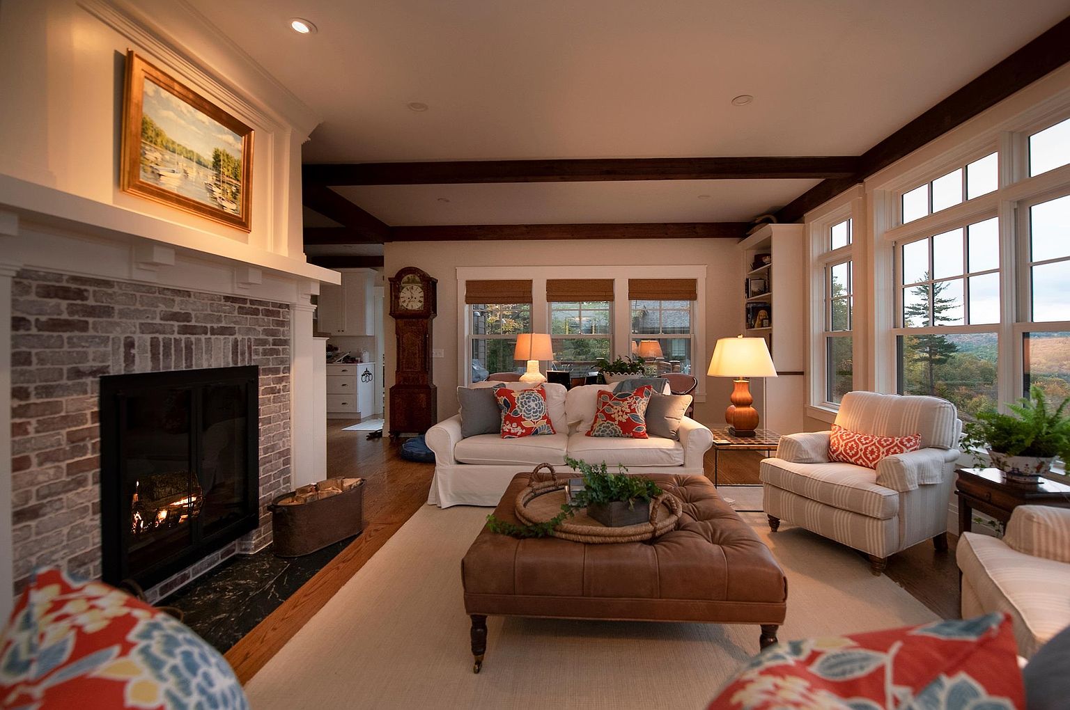 This inviting living room features a brick-faced fireplace with a wooden mantel and a framed landscape painting above. The space is anchored by a large, tufted leather ottoman and a white sofa accented with colorful floral pillows, all set upon a neutral area rug. Large windows provide ample natural light and views of the outdoors, while exposed wooden ceiling beams add a rustic, architectural touch to the room.