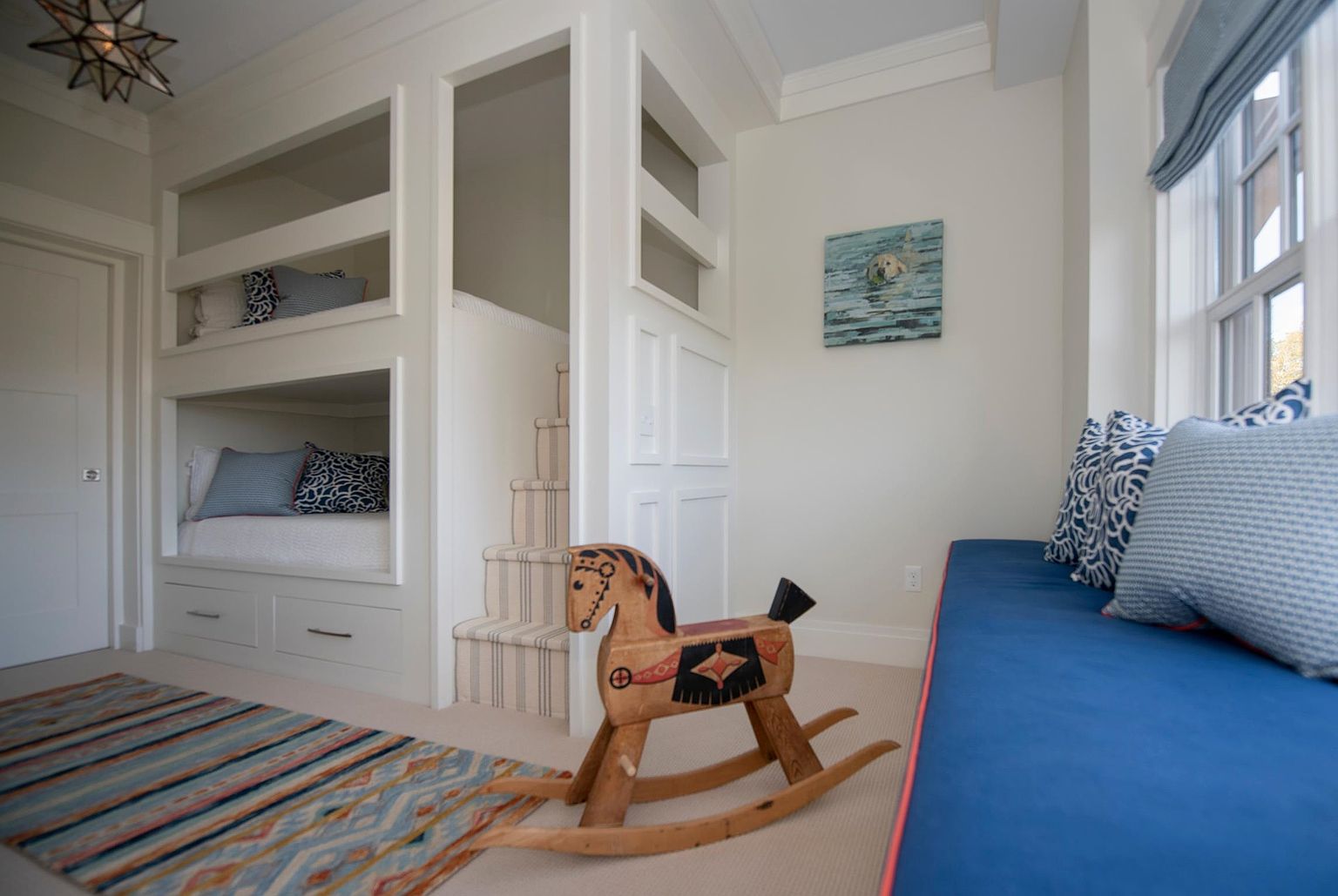 This charming children's bedroom features custom-built white bunk beds with integrated stairs and storage drawers. A vibrant, multi-colored striped rug sits on the carpeted floor near a classic wooden rocking horse, while a long blue window seat with patterned pillows provides a cozy reading nook. The room is bright and inviting, accented by a unique star-shaped pendant light and a playful painting of a dog on the wall.