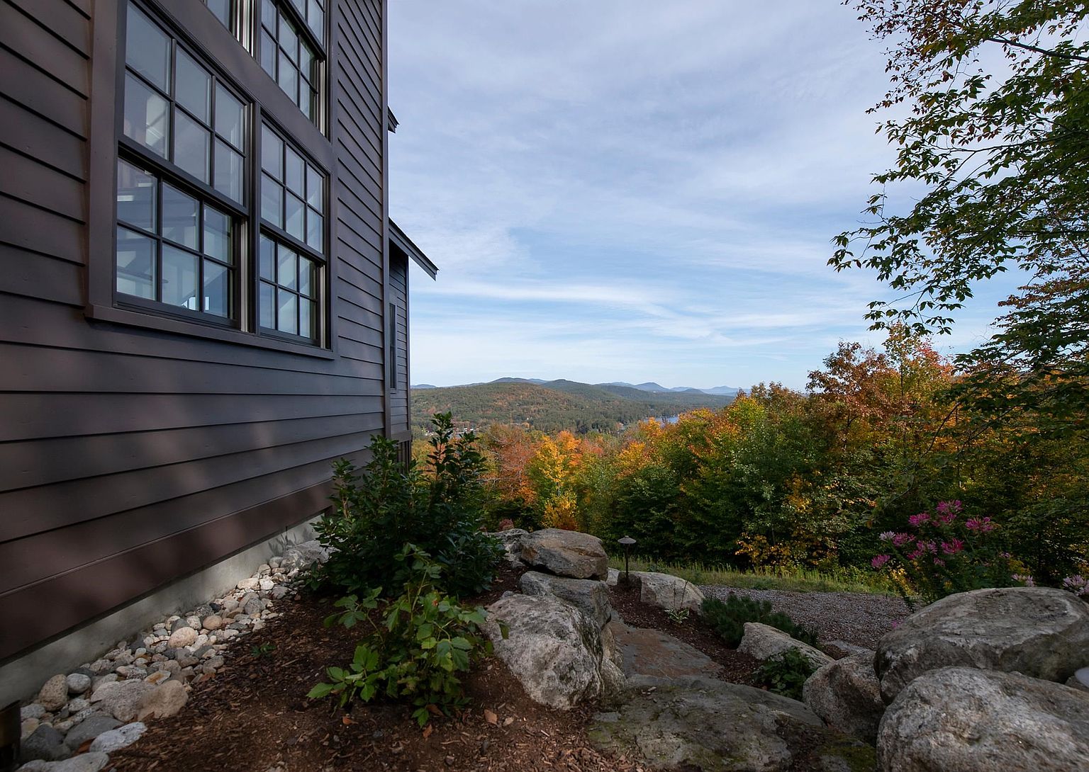 This side view of a modern home showcases dark horizontal siding and large, multi-pane windows that reflect the surrounding landscape. The house is nestled into a sloped, naturalistic landscape featuring large boulders and native plantings, leading the eye toward a scenic vista of rolling, autumn-colored hills. The perspective captures the seamless integration of the architectural structure with the rugged, wooded environment.