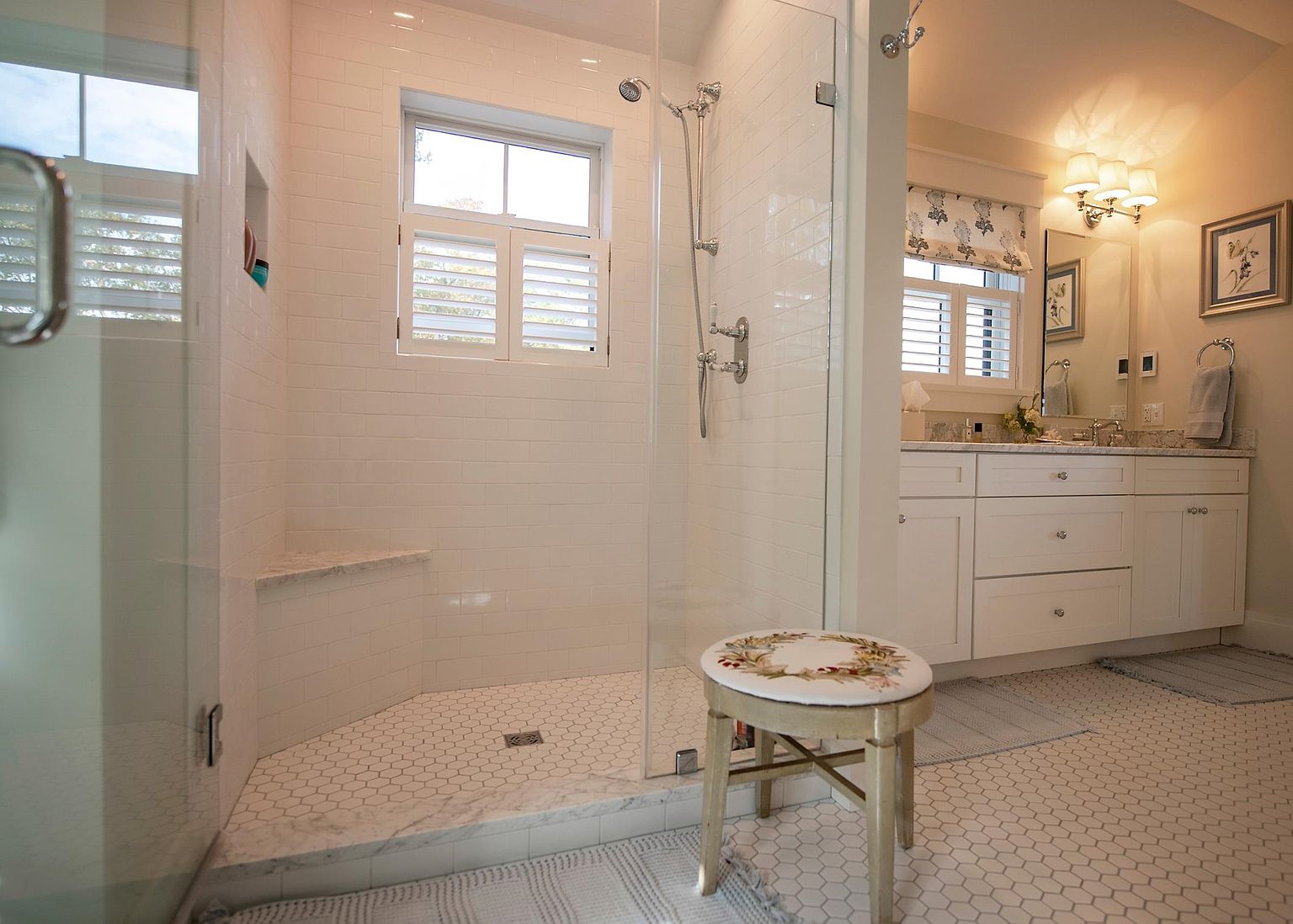 This bright and airy bathroom features a walk-in shower with white subway tile walls, a built-in stone bench, and a glass enclosure. A white vanity with a stone countertop and double sinks sits adjacent to the shower, complemented by a decorative stool in the foreground. The space is finished with a classic hexagonal tile floor, creating a clean and cohesive aesthetic.