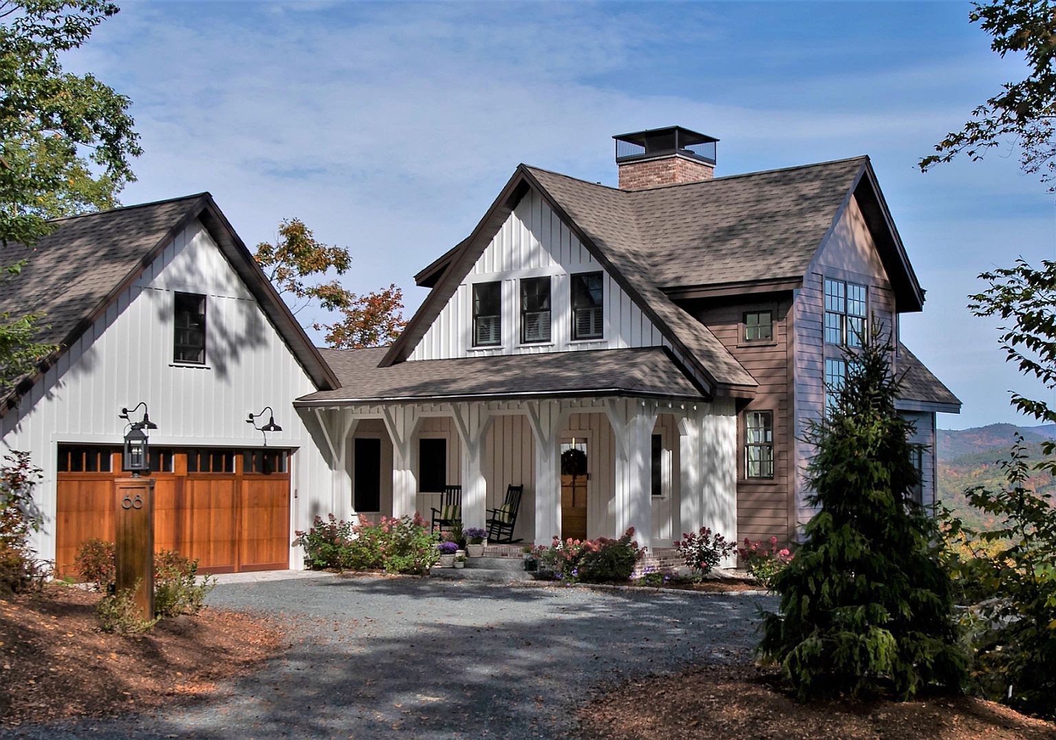 This charming farmhouse-style home features a striking combination of white vertical siding and warm-toned horizontal wood accents. The front facade is highlighted by a welcoming covered porch with decorative brackets and a gravel driveway leading to an attached garage with wooden doors. A prominent brick chimney rises from the roofline, and the surrounding landscape includes mature trees and lush garden beds, creating a serene and inviting mountain retreat atmosphere.