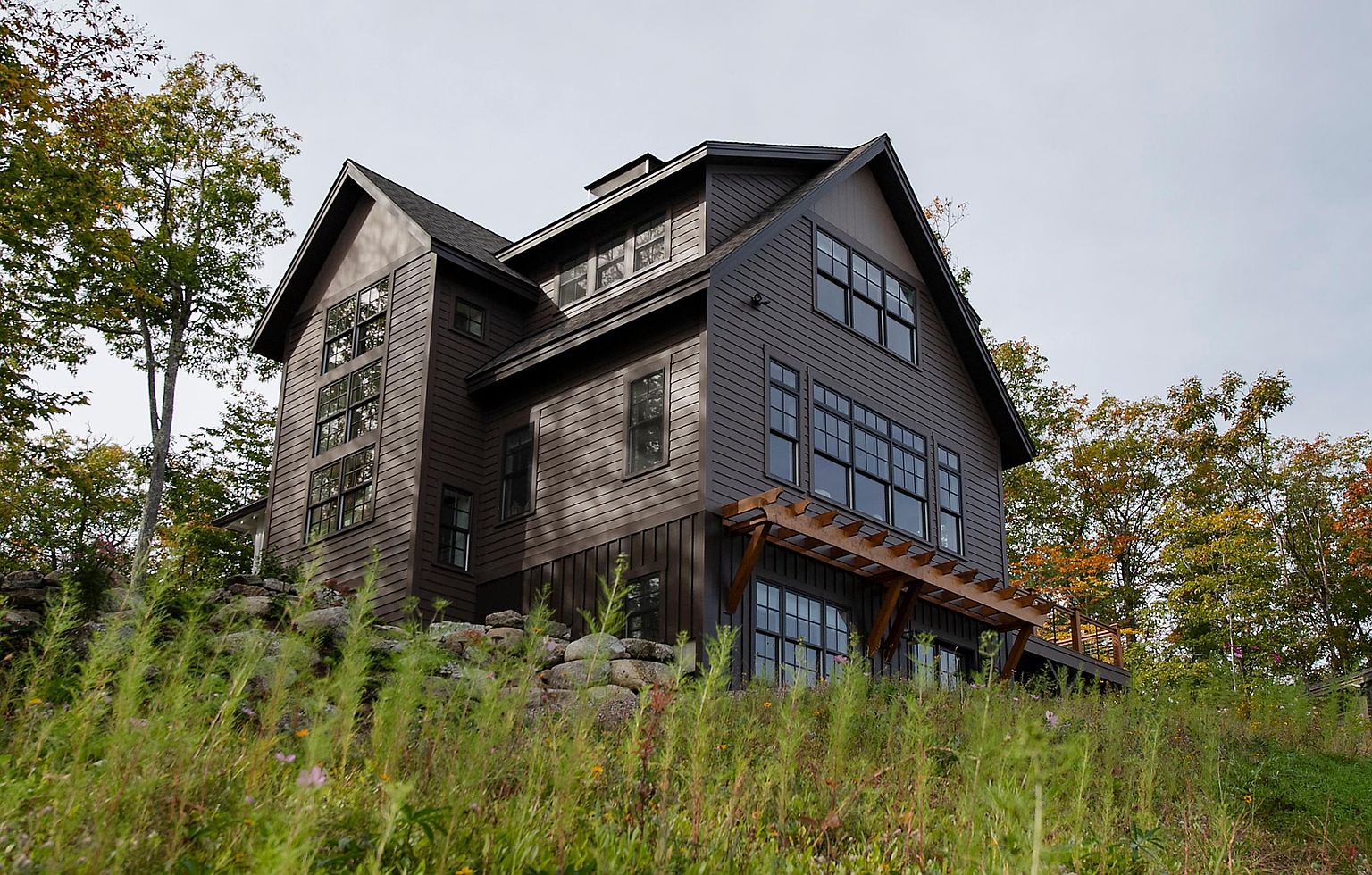 This striking multi-story home features dark horizontal siding and a complex roofline, set against a backdrop of lush, autumn-tinged trees. The structure includes a prominent wooden pergola extending over a lower-level deck, with numerous large windows that invite natural light into the interior. The house is elevated on a stone-retaining wall foundation, emphasizing its integration into the sloped, natural landscape.