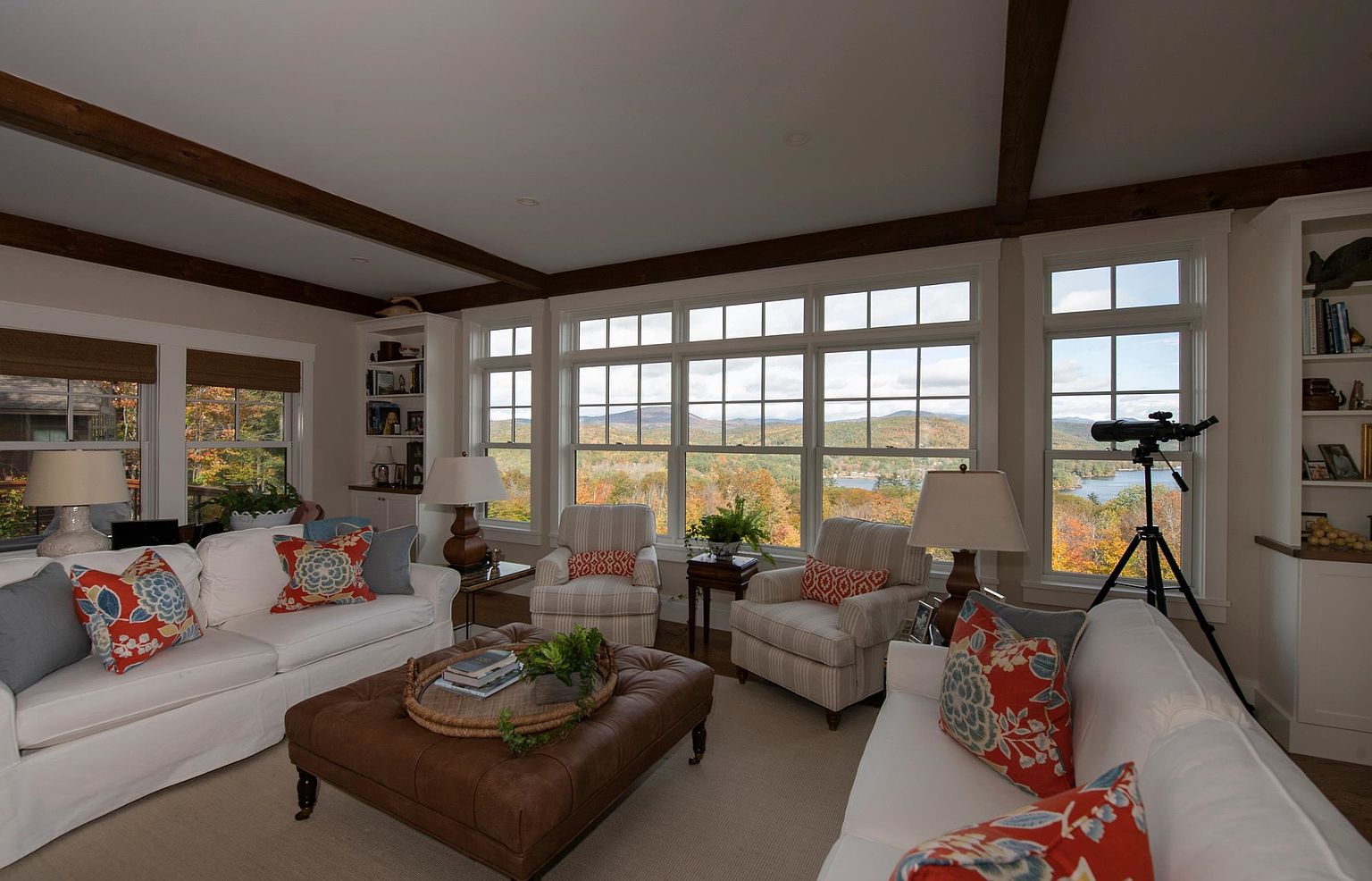 This inviting living room features a symmetrical layout with two white sofas facing each other, centered around a large tufted leather ottoman. The space is framed by expansive windows that offer a panoramic view of a scenic, tree-lined landscape and a distant lake. Exposed wooden ceiling beams and built-in shelving units add character to the room, while patterned throw pillows and warm lighting create a cozy, comfortable atmosphere.