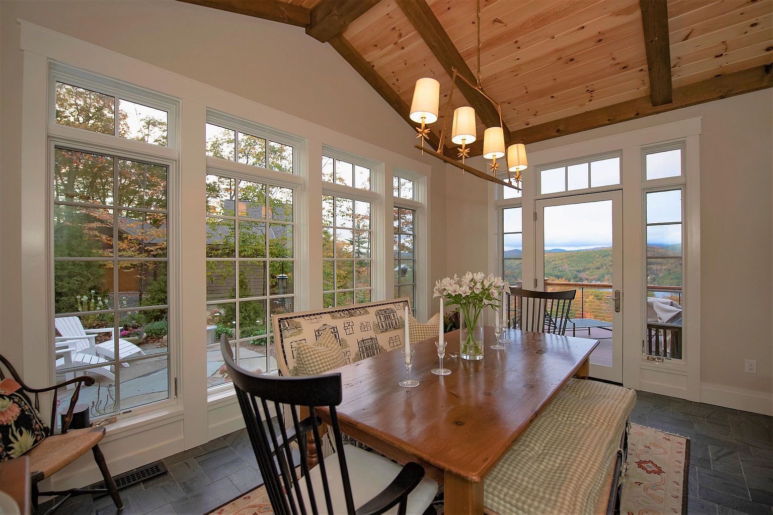 This bright dining room features a rustic wooden table surrounded by a mix of black spindle chairs and a cushioned bench, creating a cozy and inviting atmosphere. The space is defined by large windows that offer expansive views of the surrounding landscape and a wood-paneled ceiling with exposed beams. A multi-light chandelier hangs above the table, while the slate-tiled floor adds a durable and natural texture to the room.