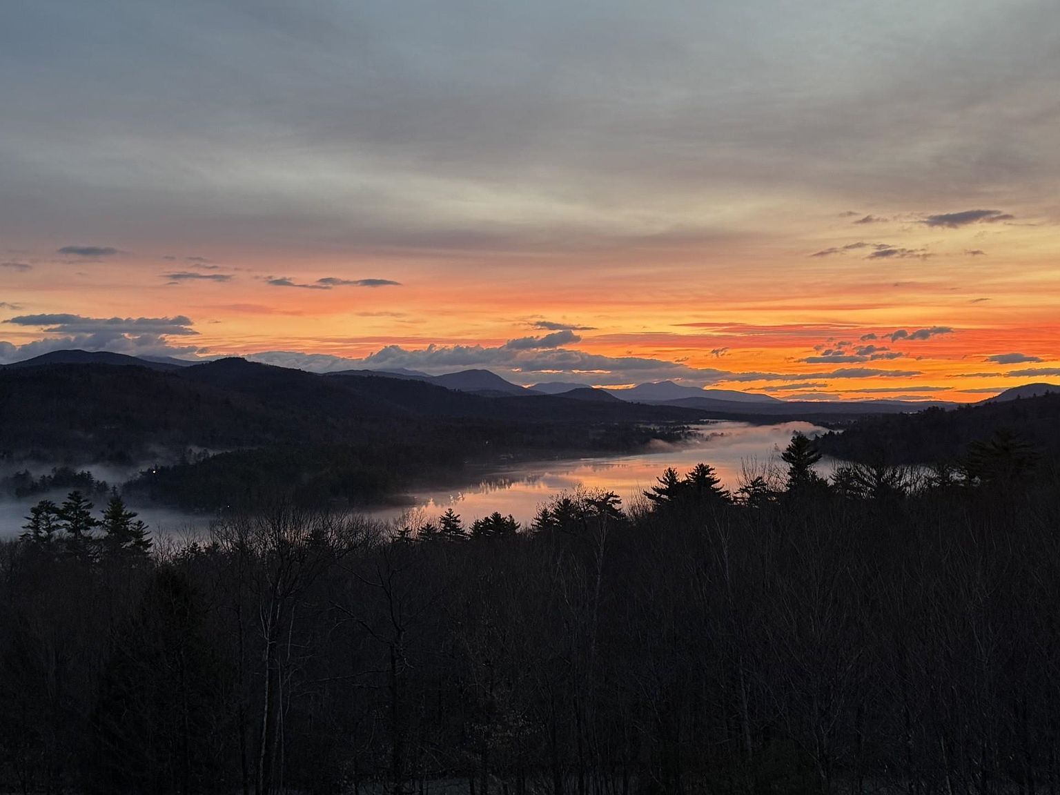 This scenic landscape view captures a breathtaking sunrise over a mist-covered lake nestled between rolling mountain ranges. In the foreground, a dense silhouette of pine trees frames the horizon, creating a dramatic contrast against the vibrant orange and purple hues of the morning sky. The perspective is elevated, suggesting a vantage point from a high-altitude property or a scenic overlook, emphasizing the tranquil and expansive natural beauty of the surrounding environment.